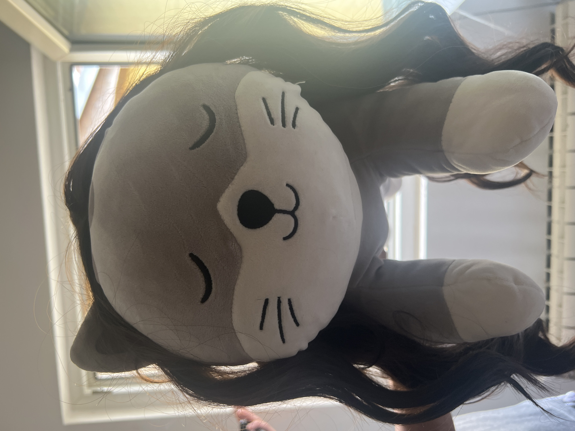 A stuffed animal, likely a cat or similar creature, is upside down and surrounded by dark brown hair. The stuffed animal has a light gray head with white accents on the muzzle, paws, and ears. It features cartoonish facial details such as closed eyes, a small nose, and whiskers. The background shows a window frame with natural light shining through. The location is Unknown, Unknown.