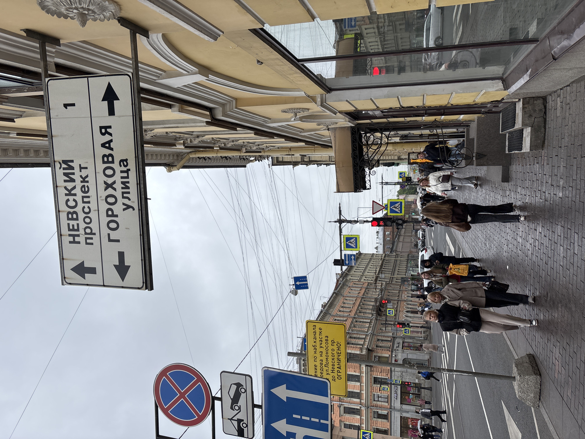 The image shows a street scene in Saint Petersburg, Russia. The location is indicated by street signs, including one with the name "НЕВСКИЙ проспект" (Nevsky Prospekt) and "ГОРОХОВАЯ улица" (Gorokhovaya Street), and another with text written in Cyrillic. There are multiple traffic lights with red lights. Several pedestrians are walking on the sidewalk, and a wheelchair is near a building. Numerous wires are strung across the sky. Road signs indicate driving prohibitions and directions. The sky is overcast.