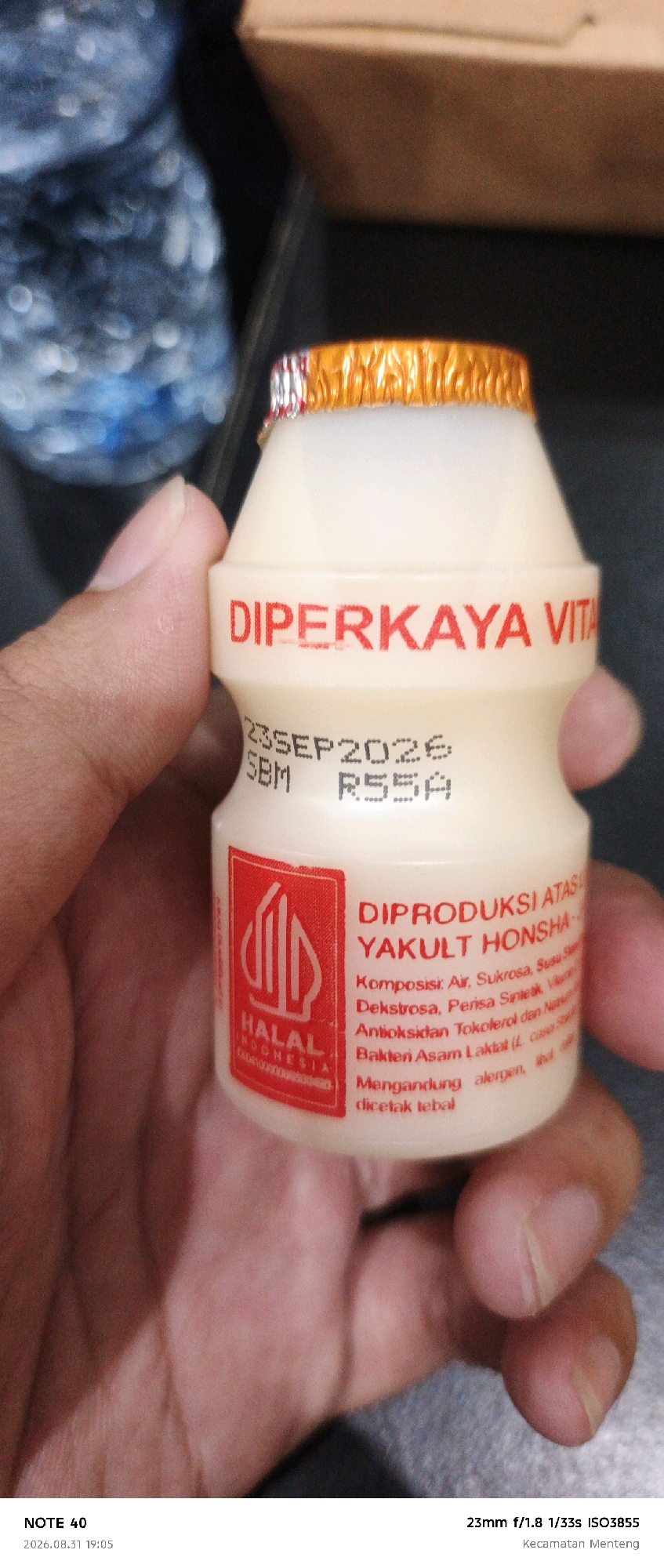 A hand holds a small bottle of Yakult Honsha drink. The bottle has a white plastic body and a foil cap with orange and silver accents. Red text on the bottle reads "DIPERKAYA VITA" at the top and "DIPRODUKSI ATAS YAKULT HONSHA" below, with a list of ingredients and "HALAL INDONESIA" in a red rectangle with a white design. A date stamp "23SEP2026 SBM R55A" is visible below the "DIPERKAYA VITA" text. The background shows a blurry blue bottle on the left and a brown cardboard box in the upper right corner. The lower part of the image contains text indicating "NOTE 40 2026.08.31 19:05" and "23mm f/1.8 1/33s ISO3855 Kecamatan Menteng". The presence of "Kecamatan Menteng" suggests the location is in Menteng District, Central Jakarta, Indonesia.