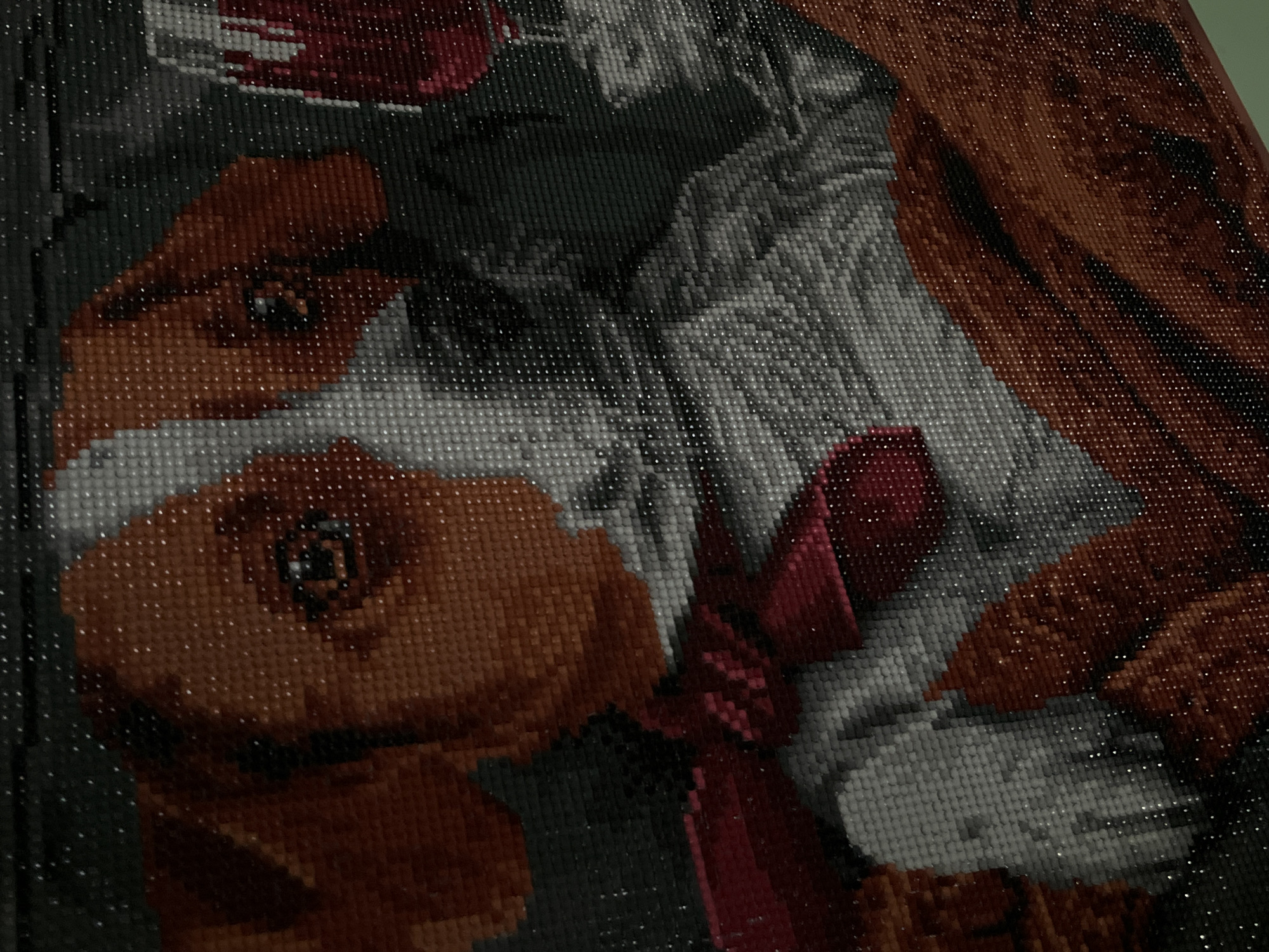 This is a close-up, high-angle, full shot of a diamond painting. The painting depicts a close-up of an animal, likely a dog, with large, expressive eyes and a reddish-brown snout and ears. The animal is wearing a red and white scarf or bandana around its neck. The background is dark and abstract, with some lighter gray and white areas that suggest fur or possibly clothing. The overall impression is one of warmth and affection. The diamond painting technique gives the image a textured, mosaic-like appearance. The lighting is somewhat dim, with a slight glare reflecting off the surface of the painting, which is typical of diamond art. No visible text is present. The context provided places this image in Lviv, Ukraine, but there are no visual cues within the image to confirm or deny this location. The activity taking place is the creation or display of the diamond art.