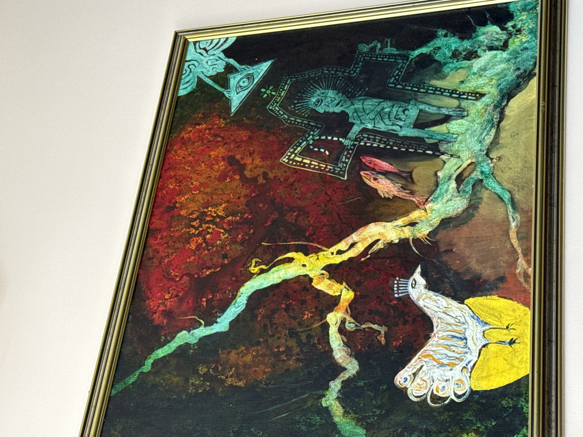 The image shows a framed painting hanging on a white wall. The painting features a complex, surreal composition with a dark background that transitions from a red, textured upper section to a greenish-black lower area. Prominent elements include a bright yellow disc on the right side with a white, stylized bird-like figure superimposed on it, an elongated, twisting shape resembling a tree trunk or vine spanning the center, and cyan, geometric shapes in the upper portion that resemble ancient glyphs. The painting is in a gold-colored frame.