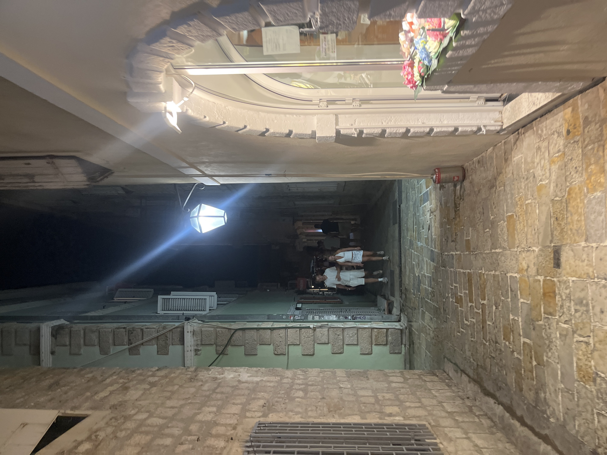 The image shows a narrow cobblestone alleyway in Budva, Montenegro, at night. A young couple, a man and a woman, are walking away from the camera towards the background. The man is wearing a white polo shirt and shorts, and the woman is wearing a white top and a patterned skirt. They are walking side-by-side, with the man's arm around the woman's waist.
On the right side of the alley, there is a building with a light-colored facade. There is a storefront with a large arched window, decorated with colorful artificial flowers. The window displays some items, but they are not clearly visible. An external light fixture is mounted above the window.
On the left side of the alley, there is a building constructed with rough-hewn stone blocks. It has a metal-grated window. The buildings on both sides of the alley are tall and close together, creating a sense of enclosure.
Overhead, a distinctive lantern-style street light is illuminating the alleyway. The cobblestone ground is wet, suggesting recent rain. In the background, other people can be seen walking in the distance, and more buildings line the alley. A red and silver cylindrical object, possibly a trash can or a cooler, is visible near the storefront on the right.