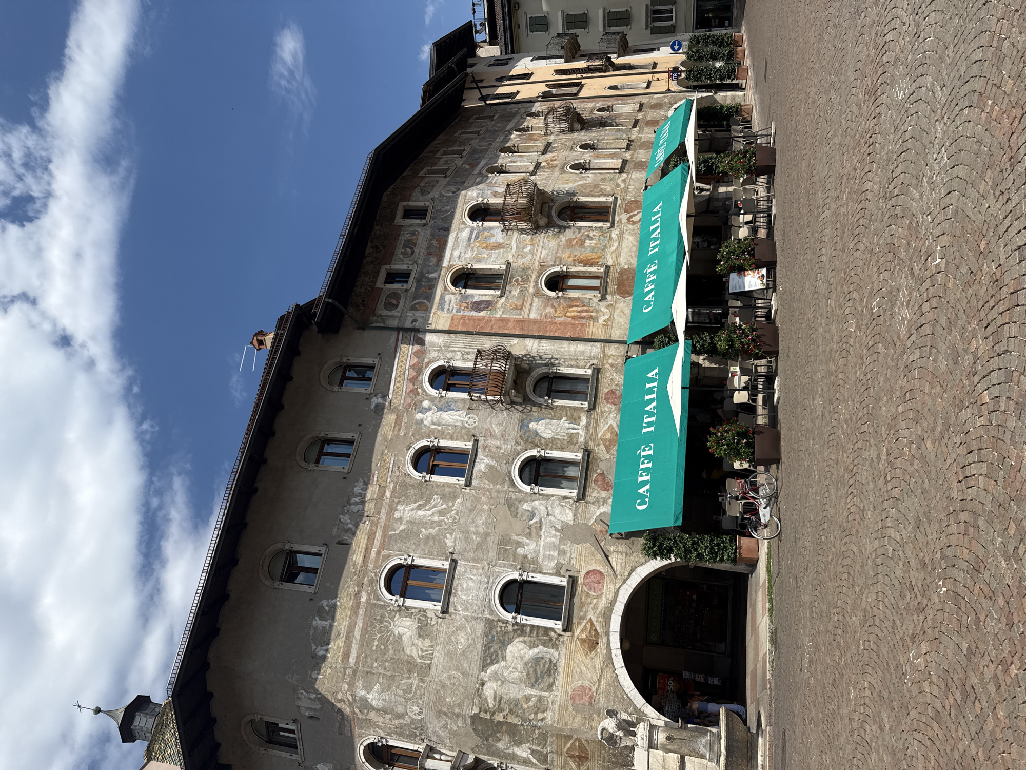 The image depicts a street view in the city of Trento, Italy, specifically in front of a building adorned with intricate frescoes. The building is a multi-story structure with arched windows and balconies. The frescoes feature scenes of figures, animals, and decorative patterns. In the foreground, a paved cobblestone square is visible. Several green awnings with the text "CAFFÈ ITALIA" are extended over outdoor seating areas for a cafe. Tables and chairs are set up under white umbrellas. A red bicycle is parked on the square. To the left of the cafe, there is an arched entrance to a shop with a sign indicating "50%". The sky above is blue with scattered white clouds. The overall scene suggests a sunny day in a historic European city.