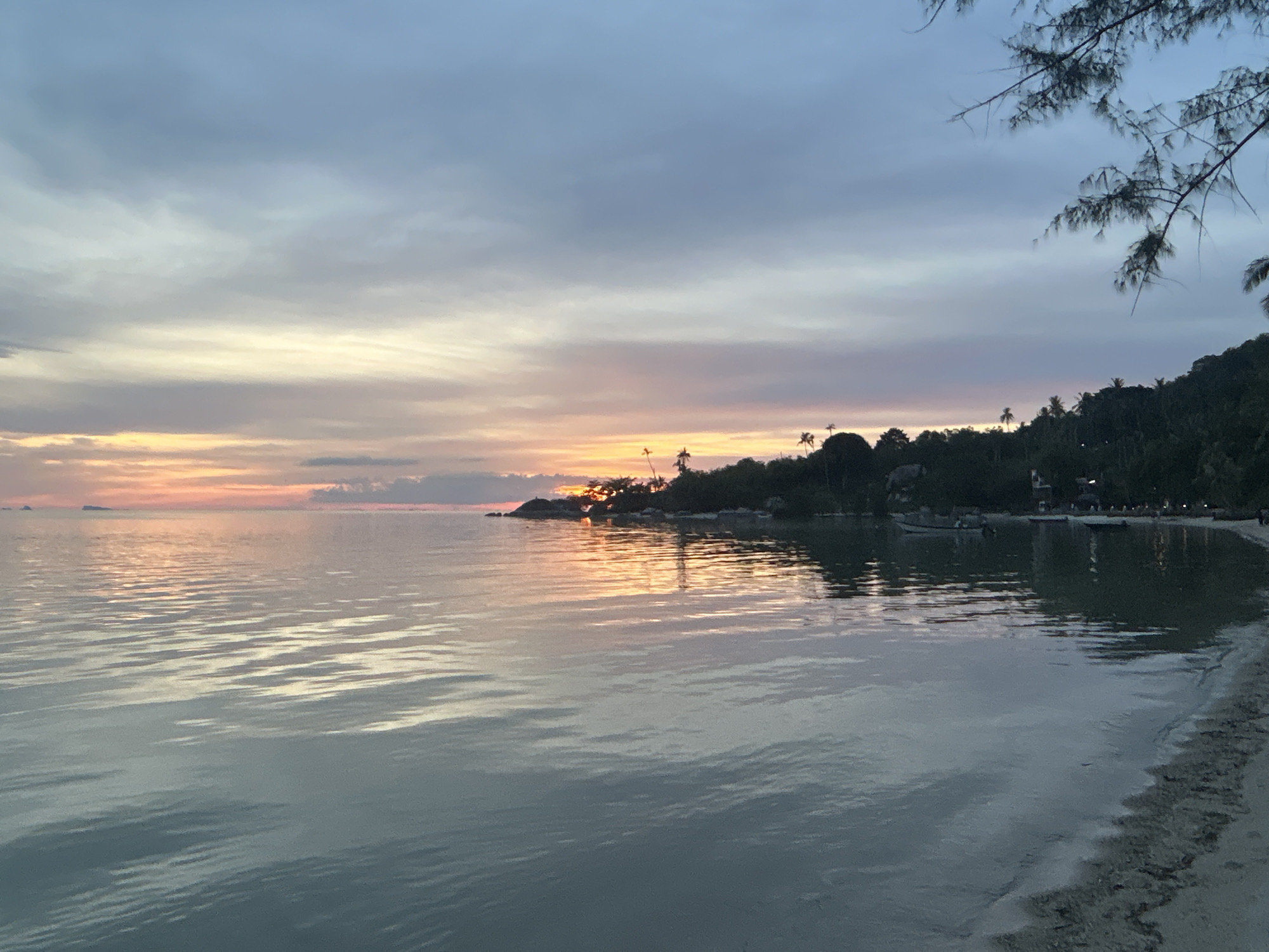 The image depicts a tranquil sunset scene at a beach in Tambon Koh Phangan, Thailand. The sky is a gradient of soft blues, purples, and oranges, with scattered clouds. The sun is setting over the horizon, casting a warm, golden light on the water and the distant treeline. The ocean is calm, with gentle ripples reflecting the colors of the sky. A sandy shoreline is visible in the foreground, with a few pieces of debris scattered along the edge. In the distance, a lush green hillside is covered with palm trees and other vegetation. There are a few small boats anchored in the shallow water near the shore. The overall atmosphere is serene and picturesque.