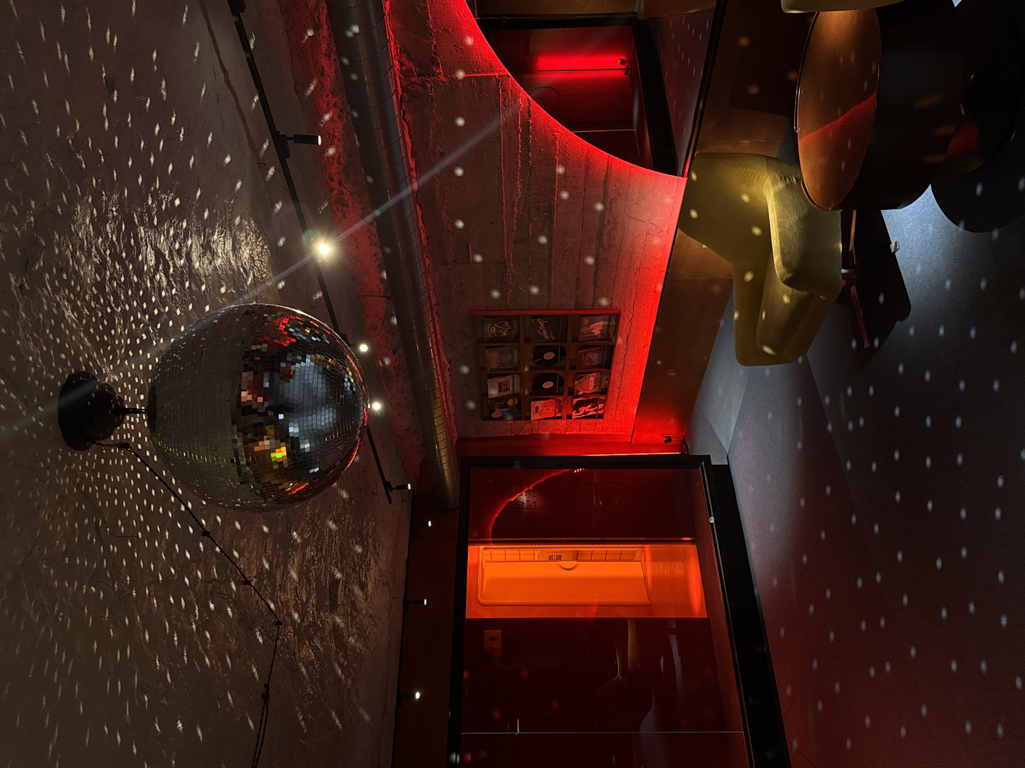 The image captures the interior of a bar or club in Yerevan, Armenia. A disco ball hangs prominently on the left, projecting small points of light onto a textured, white-painted wall. The space has a modern, industrial vibe, with exposed concrete elements highlighted by red LED lighting along the ceiling and architectural features. A set of framed vinyl record covers is visible on the wall beneath the red lighting. A doorway on the right reveals an adjacent space with orange lighting. A bar area with seating is also visible in the background. No people are directly visible in the frame. The scene suggests a vibrant nightlife atmosphere.