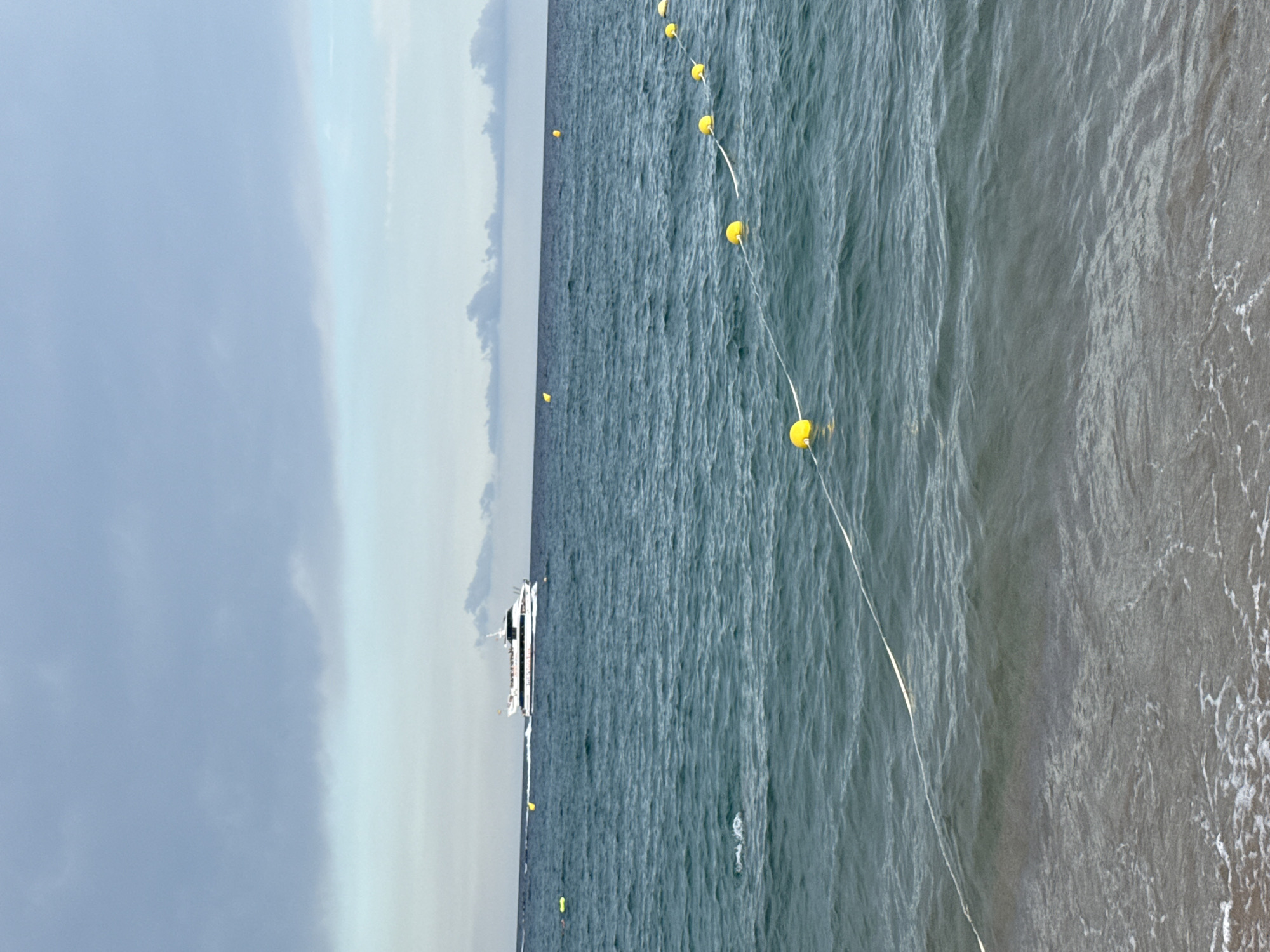 The image depicts a wide-angle view of the sea from a beach, likely in Blanes, Spain, given the context. The foreground features the gentle lapping of waves on a sandy shore, with some foam visible. A white rope with several yellow buoys is stretched across the water, delineating an area. Further out, the sea is a deep blue-green, with moderate waves. In the middle ground, a white boat with multiple decks and passengers is moving from left to right across the frame. In the background, the sky is overcast with a mix of grey and light blue clouds, with a distant, hazy horizon. The overall lighting suggests an overcast day.