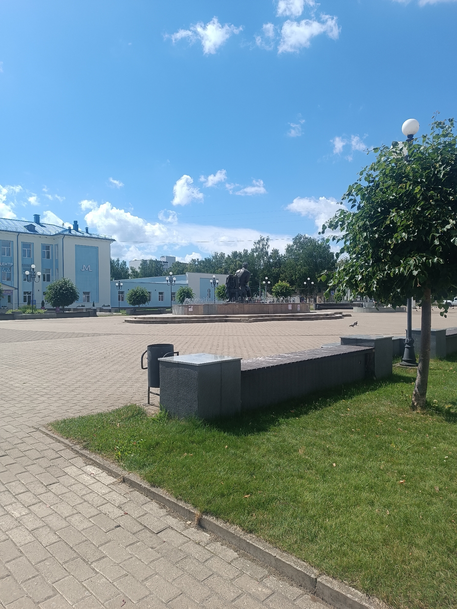 The image depicts a public square in the city of Chaikovsky, Russia, on a clear, sunny day. In the foreground, a paved walkway with interlocking gray bricks leads to a grass area with a dark gray, stone bench. A trash bin is also visible next to the bench. Further into the square, more brick paving is present, surrounding a circular fountain with water jets. Behind the fountain, a dark bronze sculpture of what appears to be a historical or mythological scene with figures and horses is situated. To the left of the fountain, a light blue building with white trim and multiple windows stands, possibly a public institution or business, with the Cyrillic word "ЦЕНТР" (Center) visible above an entrance. Several trees, including a prominent one on the right with a rounded canopy, are scattered throughout the square, providing shade. A lamppost with a globe-shaped light fixture is also visible next to the tree on the right. The sky is a vibrant blue with scattered white cumulus clouds. The scene is tranquil and appears to be a well-maintained public space.