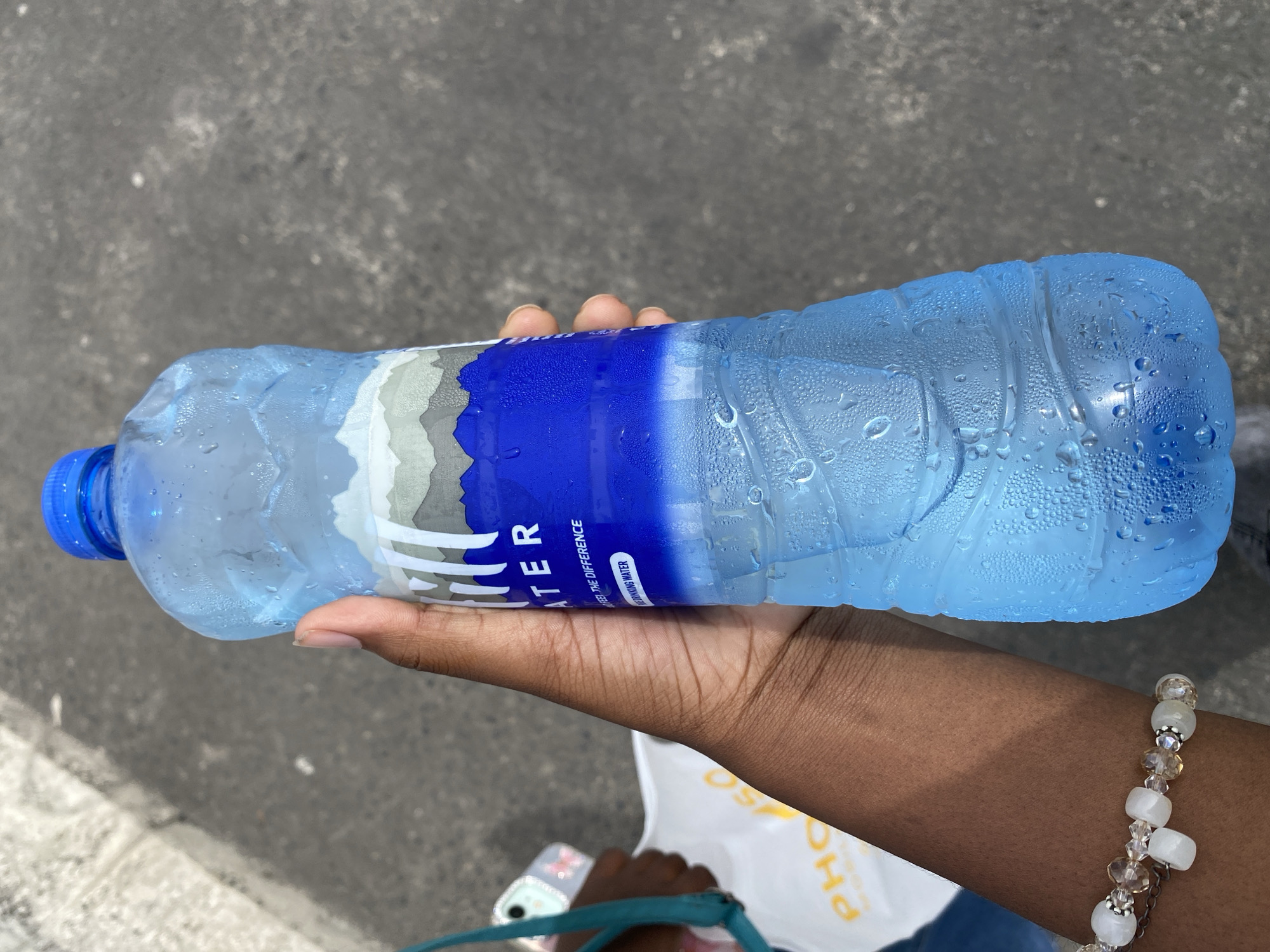 A close-up, high-angle view depicts a dark-skinned hand holding a clear plastic bottle of water outdoors. The bottle, featuring a blue screw-top cap, is visibly covered in condensation droplets, indicating a cold temperature. Its label is white, gray, and blue, with a mountain-like graphic and the text "HILL WATER," "FEEL THE DIFFERENCE," and "DRINKING WATER." The hand gripping the bottle wears a beaded bracelet composed of white and clear faceted beads. In the blurred background, to the left, a portion of another dark-skinned arm is visible, holding a white shopping bag with partially obscured yellow text, possibly "PHO...SO." A smartphone with a clear, possibly butterfly-patterned case and a teal strap is also visible behind the bottle-holding hand. The ground surface is dark gray asphalt or concrete, with a lighter gray curb or edge visible in the upper left. The scene is depicted in Dar es-Salaam, Tanzania, on an unspecified street or paved outdoor area under bright daylight.