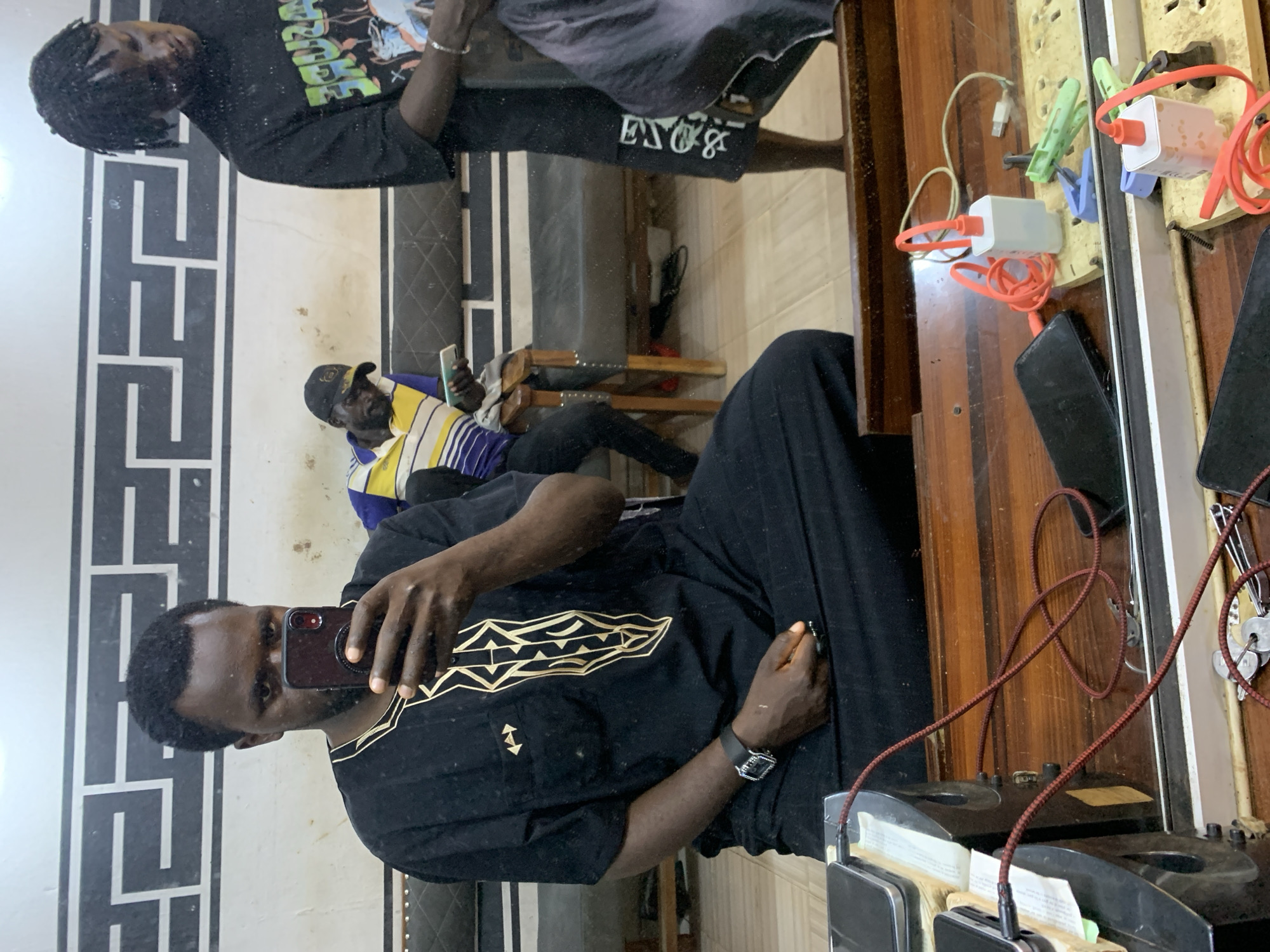 The media file is a vertical, eye-level, selfie-style shot taken in a room that appears to be a barbershop in Jos, Nigeria. The main subject is a man wearing a black garment with gold embellishments and a watch on his left wrist. He is holding a phone in his right hand, capturing his reflection in a large mirror that dominates the scene.
In the reflection, another person is visible sitting in a barber's chair, wearing a striped shirt and a cap. He is looking at the camera. The background of the mirror shows wooden shelves with various objects, including electrical chargers, cables, and some keys. The overall setting suggests a functional, possibly busy, barbershop environment. The lighting appears to be artificial, indicating it could be any time of day. There is no visible text in the image.