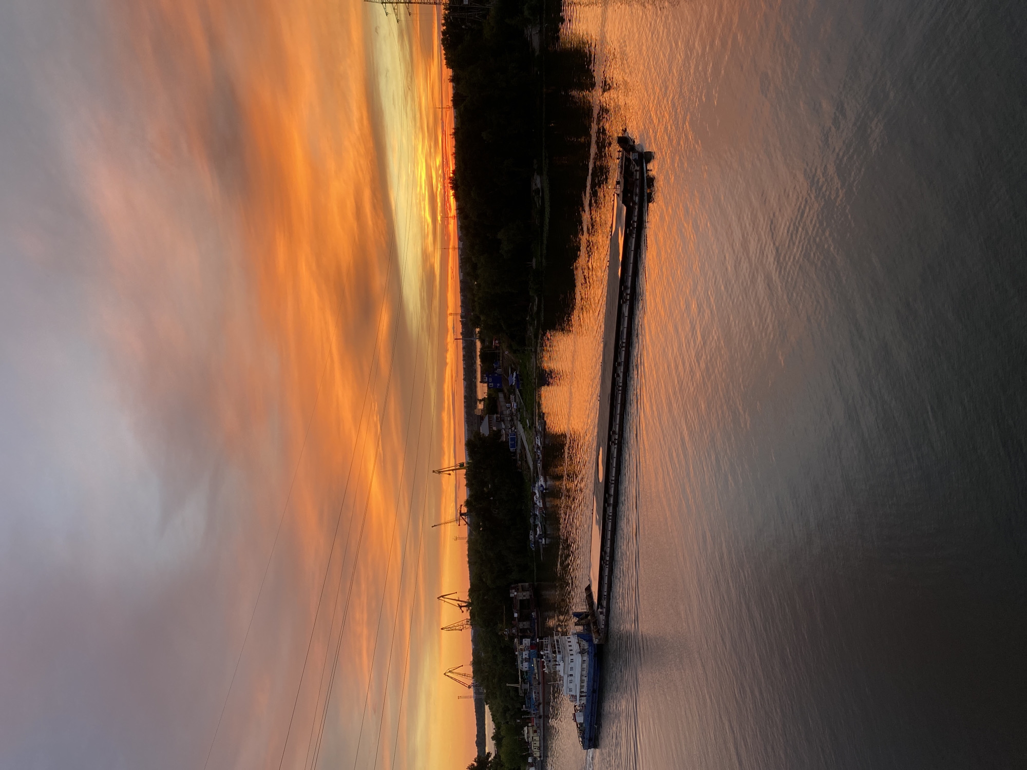 The image captures a picturesque sunset over a wide river, viewed from above. The sky is ablaze with vibrant orange and yellow hues, painted across streaky clouds, creating a dramatic and warm atmosphere. The water reflects this fiery sky, shimmering with a coppery glow, while also showing ripples from the movement of a barge.
The barge itself is a prominent object in the scene, a long, dark vessel moving down the river. It appears to be a cargo barge, possibly a push barge, with a small structure at its stern. Along the riverbank, a dense line of dark trees creates a stark contrast against the bright sky. Scattered among the trees are glimpses of human activity: buildings, structures, and several tall industrial cranes, suggesting a port or industrial area. The presence of power lines stretching across the sky also indicates proximity to human infrastructure.
The time of day is clearly sunset, given the colors in the sky and the lighting on the water. The weather appears clear, with the clouds providing a canvas for the sunset's spectacle. The location is Novokuybyshevsk, Russia, which aligns with the industrial and riverine landscape depicted. The overall mood is one of serene beauty and quiet industry, a moment where nature's grandeur meets human endeavors. There is no visible text in the image.
