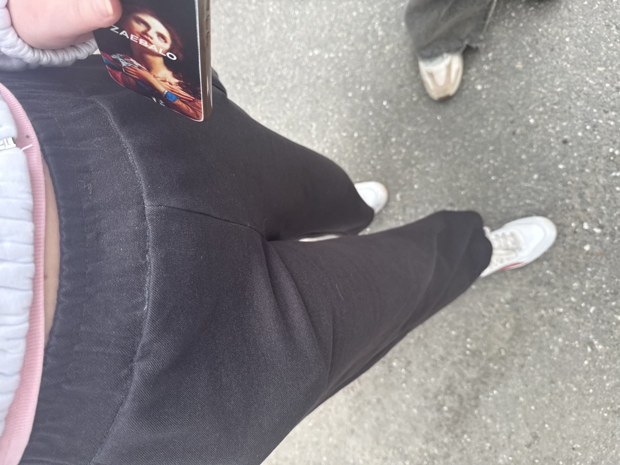 The image shows a close-up view of a person's lower body, from the waist down, as they stand on a paved surface. The person is wearing black sweatpants and white sneakers. Their left leg is bent at the knee, and their right leg is mostly obscured. In their right hand, they are holding a card with an image of a person on it. The paved surface appears to be asphalt. The image does not contain any discernible landmarks or features that would allow for a precise location within the city of Perm, Russia.