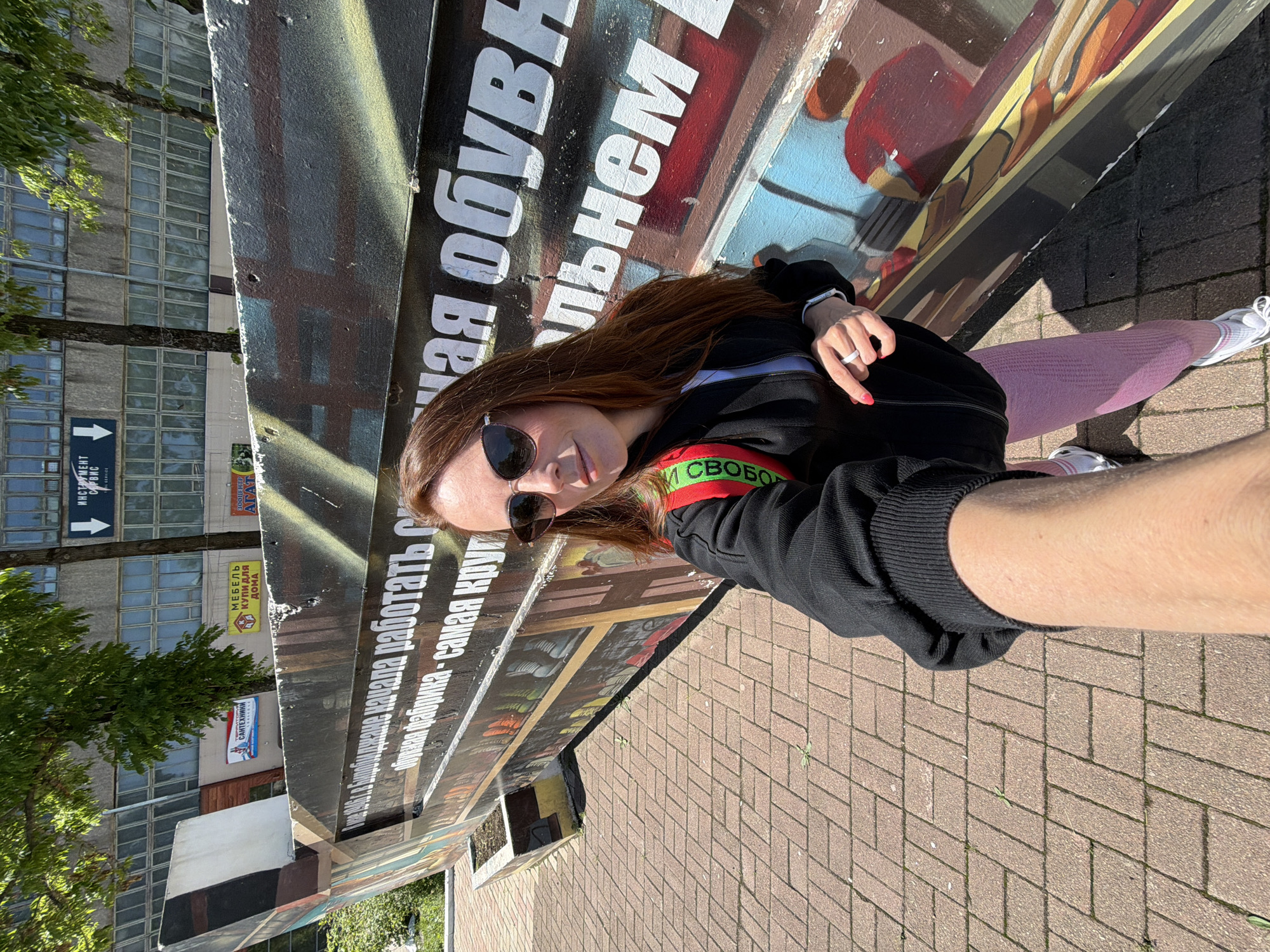 In Birobidzhan, Russia, a woman with long auburn hair and sunglasses takes a selfie in front of a mural on a sunny day. She is wearing a black zip-up jacket over a white top, pink leggings, and white athletic shoes. A red and green strap is visible over her shoulder. The mural features large white Cyrillic text on a dark background, depicting a historical narrative about a shoe factory. The ground is paved with reddish-brown bricks. Behind the mural, a building with numerous windows is visible, along with trees and signs.