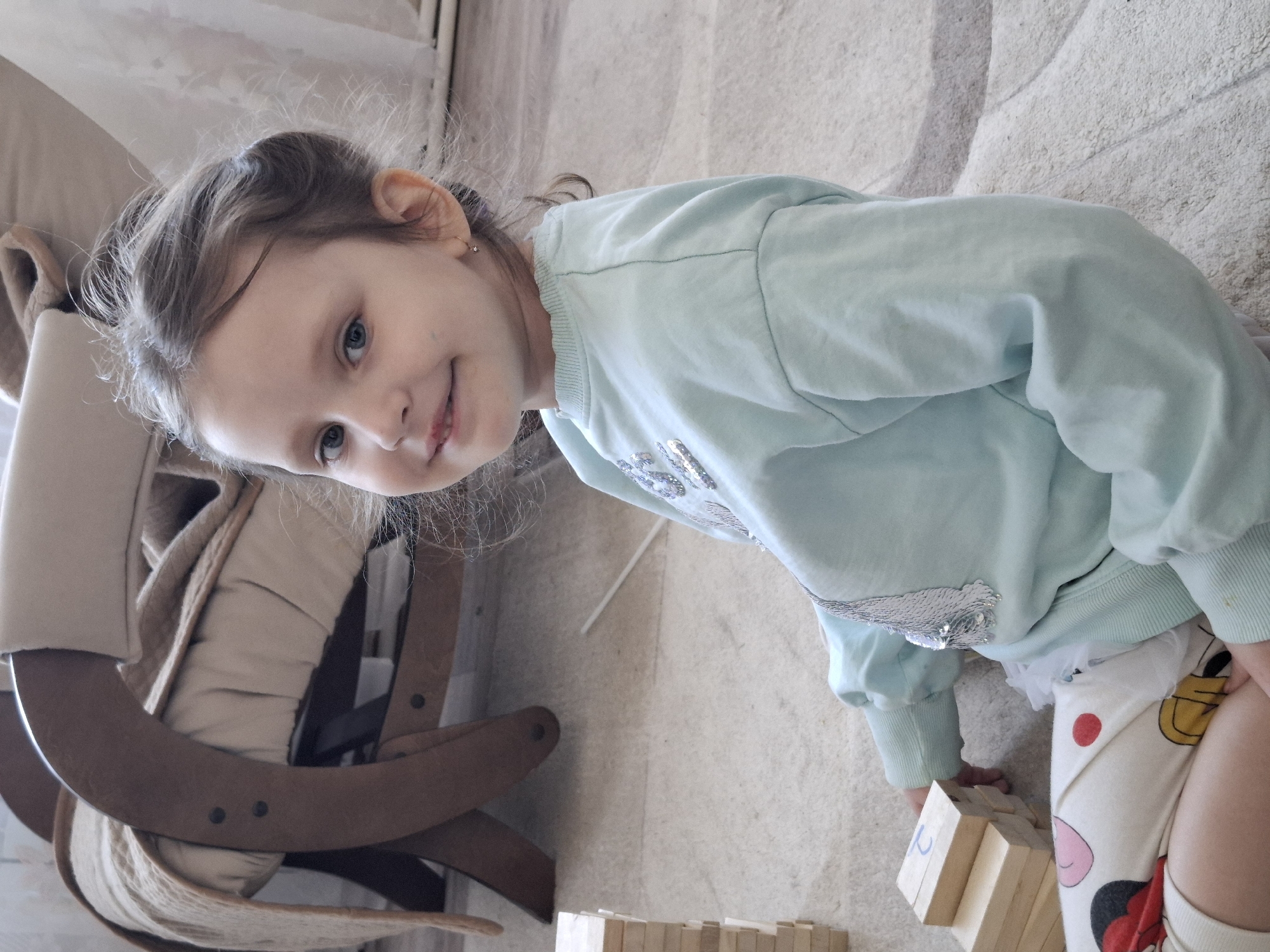 A young girl is playing with wooden blocks on the floor, next to a wooden chair with a beige cushion. She is wearing a light blue sweatshirt with a sequin design and patterned pants. The location appears to be indoors, possibly a home, but the city of Chaikovsky, Russia cannot be verified from the image.