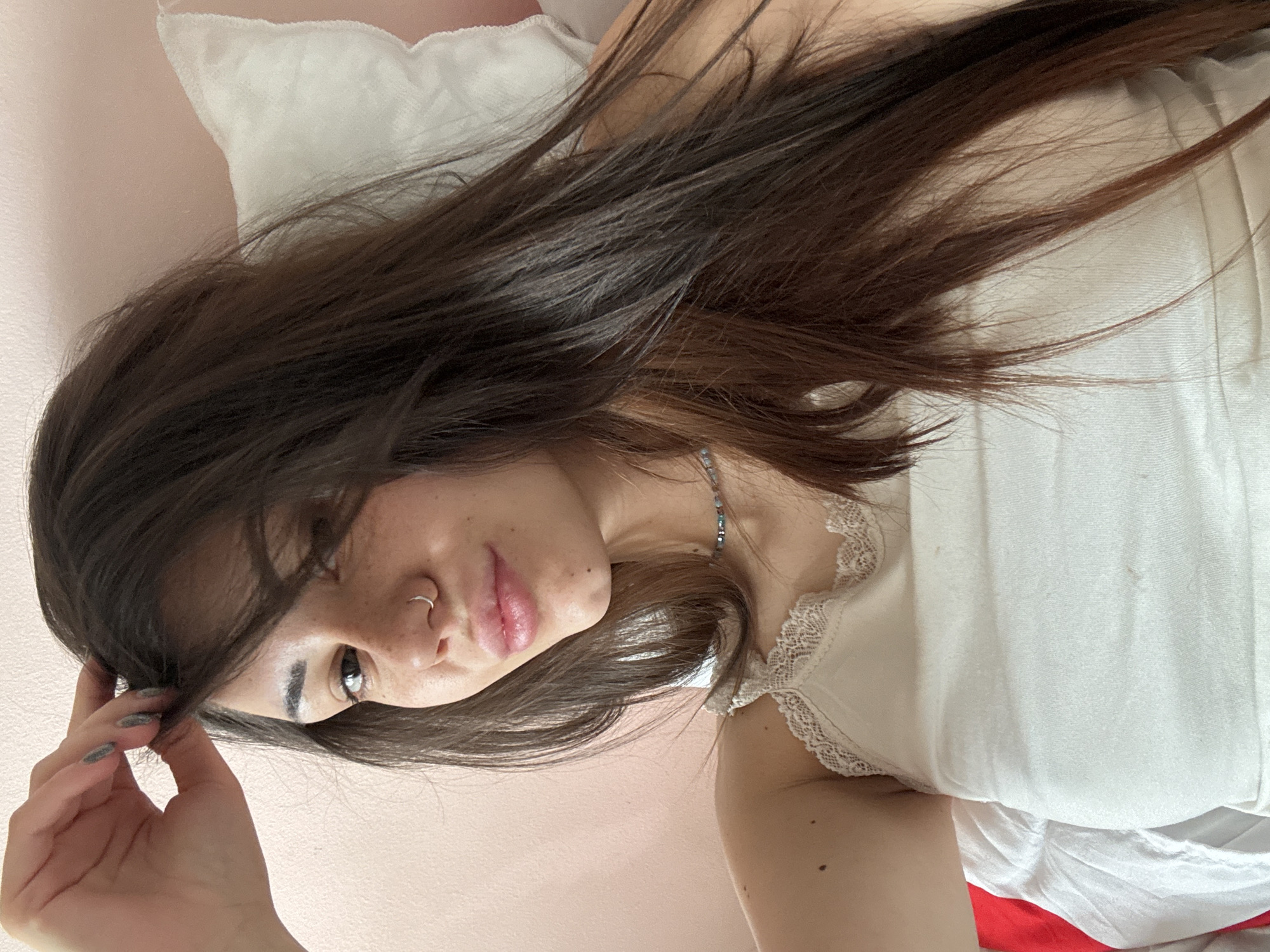 A woman is lying on a white pillow and sheet with a red accent, likely in a bedroom with pink walls. She has long brown hair, fair skin, and a nose ring. She is wearing a white top with lace trim and a beaded necklace. She is holding her hair with her left hand, which has grey nail polish. The location in the city of Sofia, Bulgaria cannot be determined from this image.