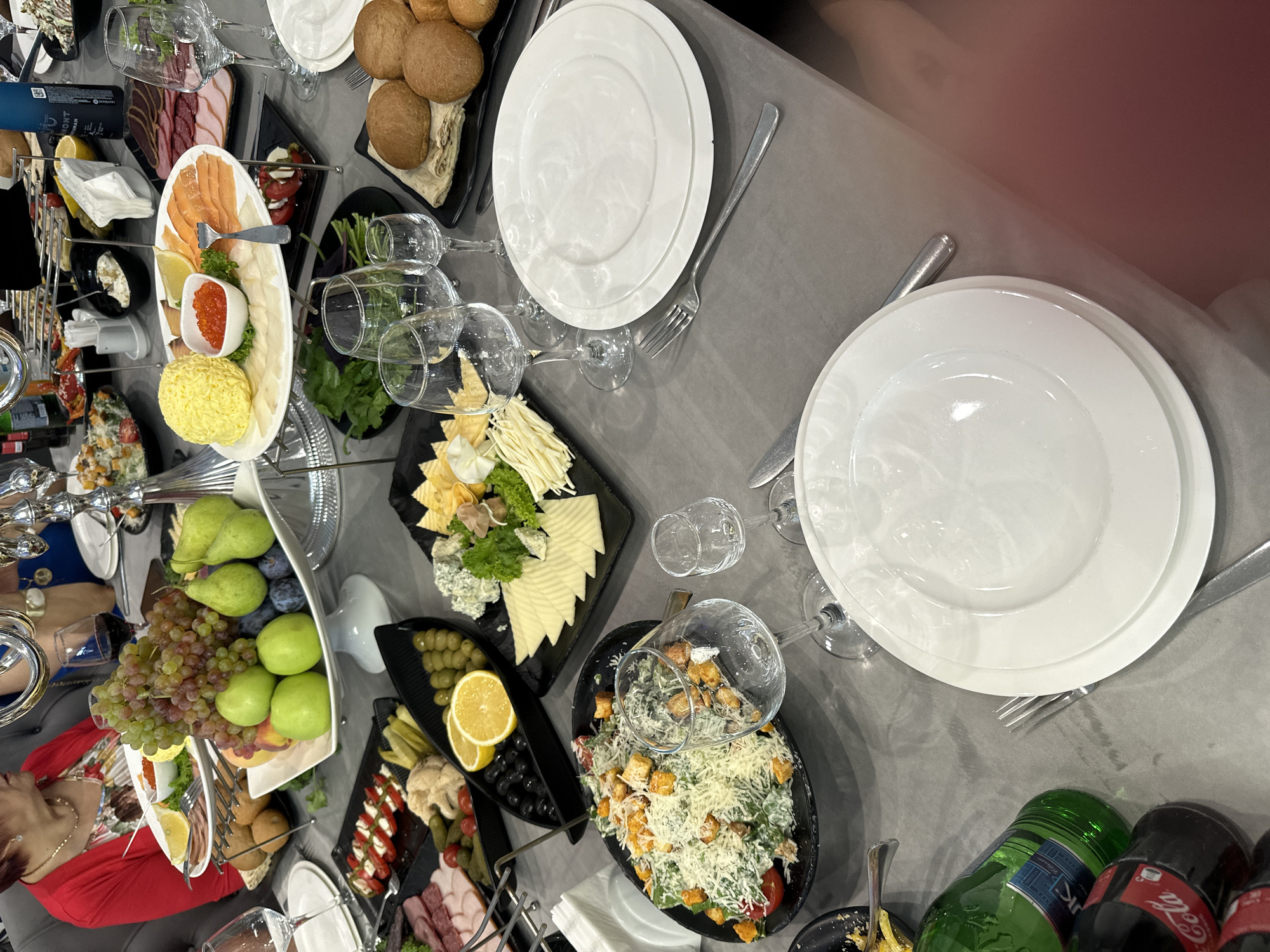 This image depicts a well-laid table with a variety of food and drinks, suggesting a celebratory meal or gathering. The setting appears to be indoors, likely a restaurant or dining room, with a grey tablecloth covering the table.
Several dishes are visible, including a large platter of assorted fruits such as grapes, apples, and pears, arranged in a decorative white bowl. Adjacent to this, there's a plate with sliced salmon, red caviar, and what appears to be cheese or butter. Another plate holds a Caesar salad topped with croutons and grated cheese. There are also small plates with olives, pickles, sliced tomatoes with mozzarella, and various types of cured meats and bread.
Several wine glasses and water glasses are placed around the table, some filled with red wine. Bottles of Coca-Cola and a green bottle, possibly soda or water, are also present. Two individuals are partially visible in the background, one wearing a red top and the other a blue top, suggesting they are attendees of the meal. The overall impression is one of abundance and a convivial atmosphere. There is no information available to confirm the location in the city of Parakar, Armenia.