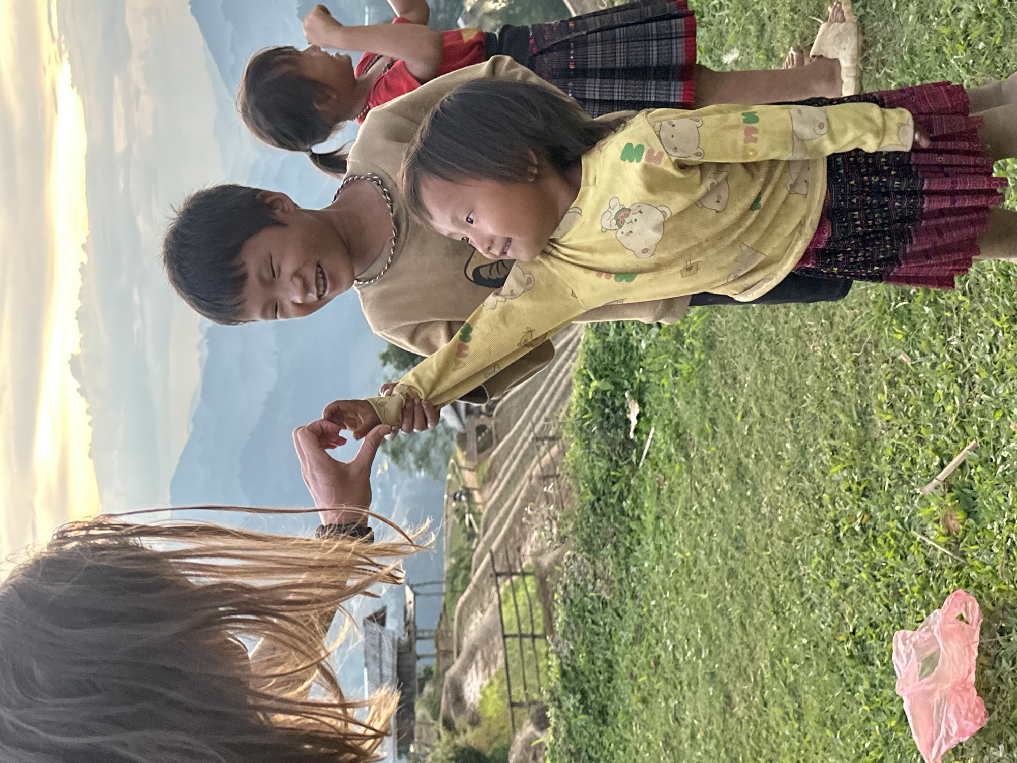 This image depicts an outdoor interaction between an adult and three children in the mountainous region of Mù Cang Chải, Vietnam, during the golden hour of late afternoon or early evening.
In the foreground, the back of an adult's head with long, light-brown hair is visible. Their hand reaches out to grasp the hand of a young girl, together forming a heart shape. The girl wears a yellow long-sleeved shirt featuring cartoon bears and the letters "MMO," paired with a patterned, dark red and black traditional skirt. She looks towards the adult's hand with a soft smile.
To her left stands a boy in a beige long-sleeved shirt and a metallic chain-link necklace, smiling broadly with his eyes closed. Behind him, another younger girl in a red shirt and a plaid skirt is partially visible, looking away. The children are barefoot, and their attire suggests traditional ethnic clothing.
The subjects are positioned on a grassy slope, with a grand backdrop of hazy blue mountains and valleys. Golden light from the setting or rising sun illuminates the distant sky and peaks. Below the children, structures resembling steps or terraced pathways descend into the landscape. A pink plastic bag lies on the grass to the right. The scene conveys warmth, connection, and joy in a scenic rural setting.