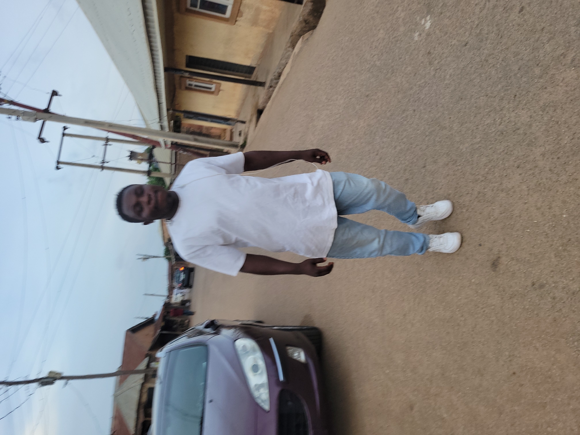 A man stands on an asphalt street next to a purple car. He is wearing a white shirt, light blue jeans, and white sneakers. The background features buildings, utility poles, and other cars. The location is Jos, Nigeria.