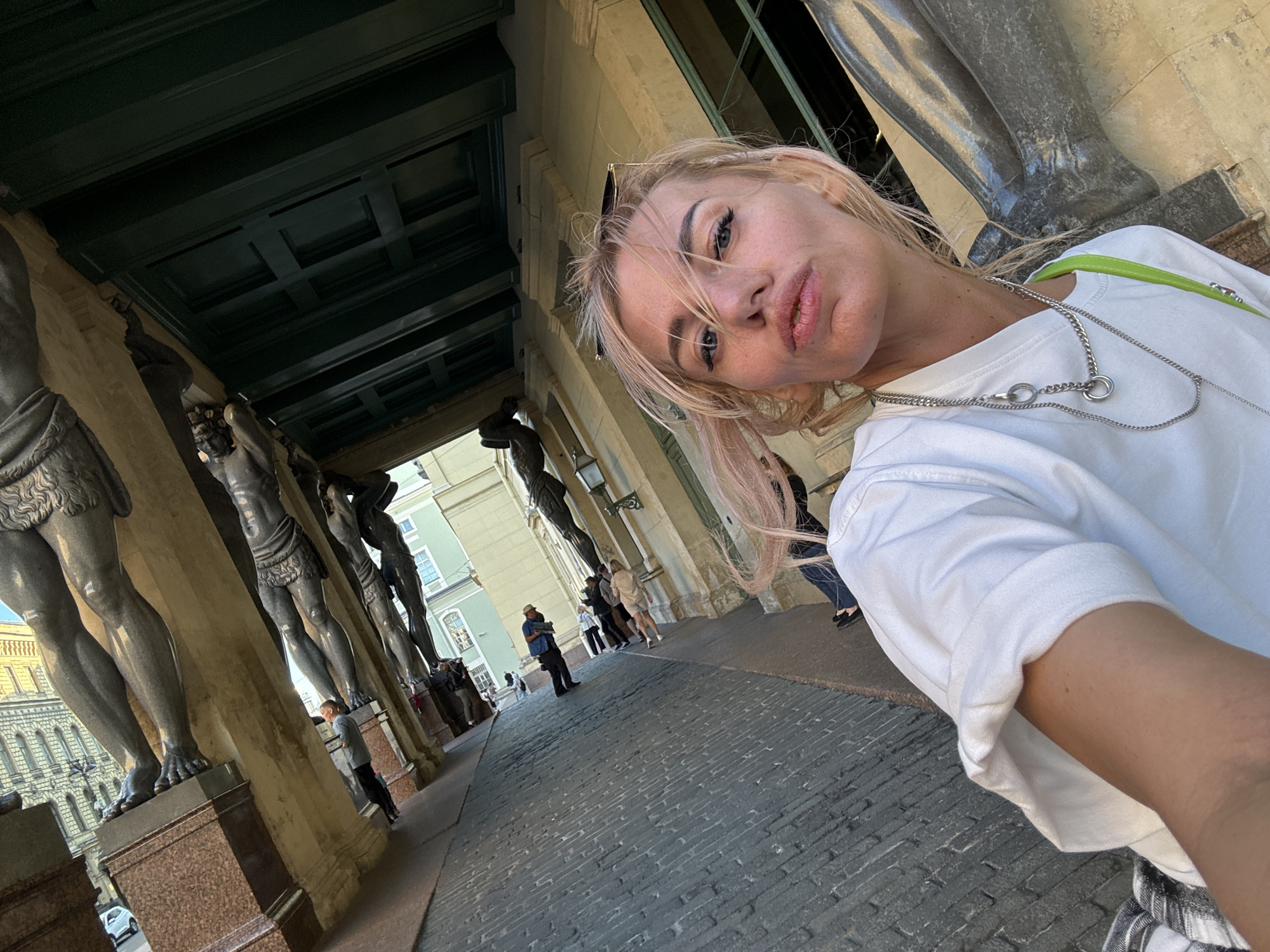 This media file captures a woman taking a selfie outdoors in front of a historic building with a colonnade. The woman, with blonde hair and makeup, is wearing a white t-shirt and a necklace. Behind her, the scene features large, stone statues of men holding up the building's structure, a common architectural element in classical design. The ground is paved with cobblestones. Several other people are visible in the background, suggesting it's a public space with some foot traffic. The lighting suggests it is daytime, with the sun casting shadows. The woman's facial expression is a pout, perhaps playful or posed for the selfie. The overall setting implies a tourist or historical site, consistent with the location context of Saint Petersburg, Russia, known for its grand architecture and sculptures.