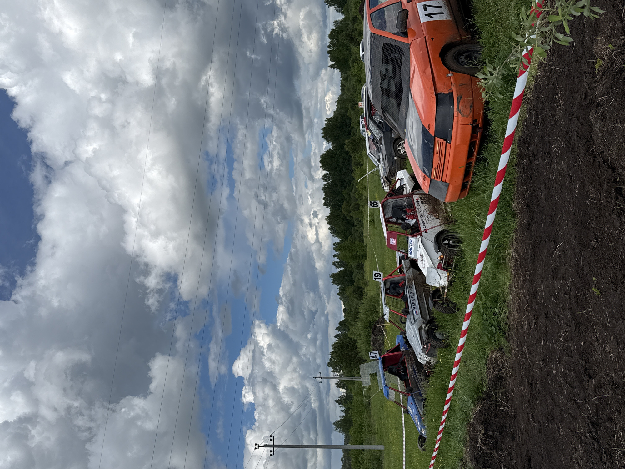 The image depicts a scene in Votkinsk, Russia, featuring a row of modified vehicles parked on a grassy field next to a dirt track. The vehicles appear to be prepared for some type of racing or off-road event. The vehicles have roll cages and competition numbers, with one prominently displaying the number 17. The surroundings consist of trees, a field, and power lines against a cloudy sky. A red and white striped barrier marks the edge of the track.