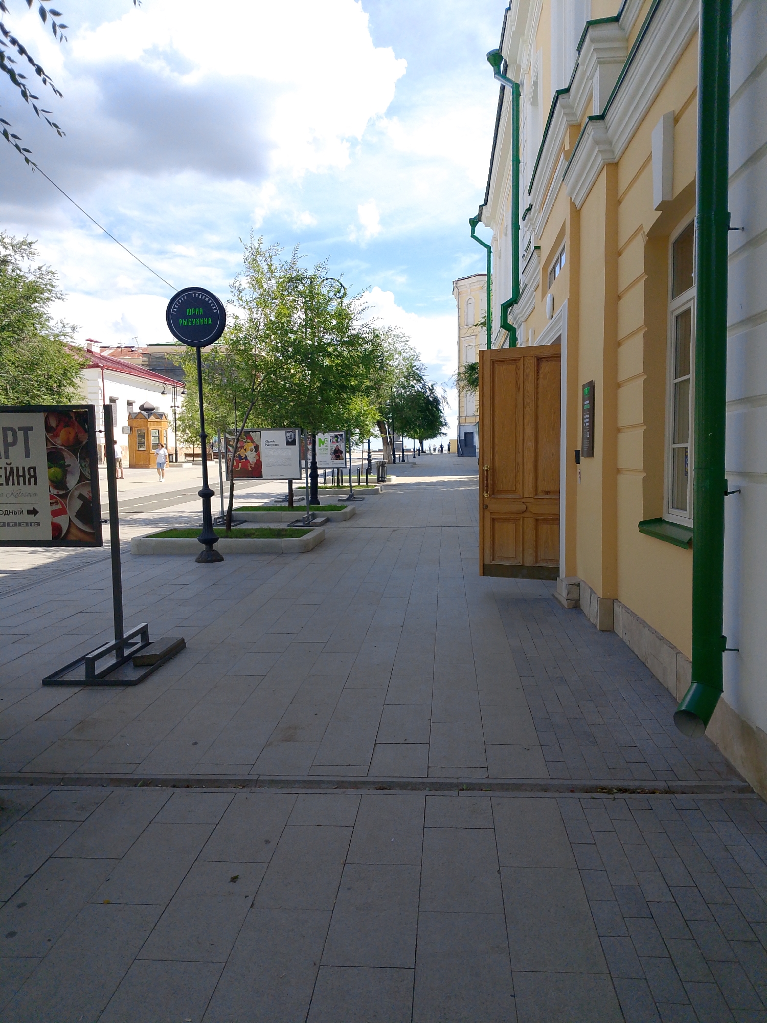 The image shows a street in Orenburg, Russia, with a pedestrian sidewalk lined by trees and a building. On the left, there is a sign with Cyrillic text reading "АРТ" and "ЕЙНЯ", indicating an art-related exhibition. A circular sign with green text is mounted on a pole. In the background, a building with yellow walls and white trim is visible, featuring an open wooden door and a green downspout. The sidewalk is paved with grey stone tiles. Several art display stands with posters are positioned along the sidewalk. The sky is cloudy with patches of blue. The overall scene depicts a public space possibly used for outdoor exhibitions.