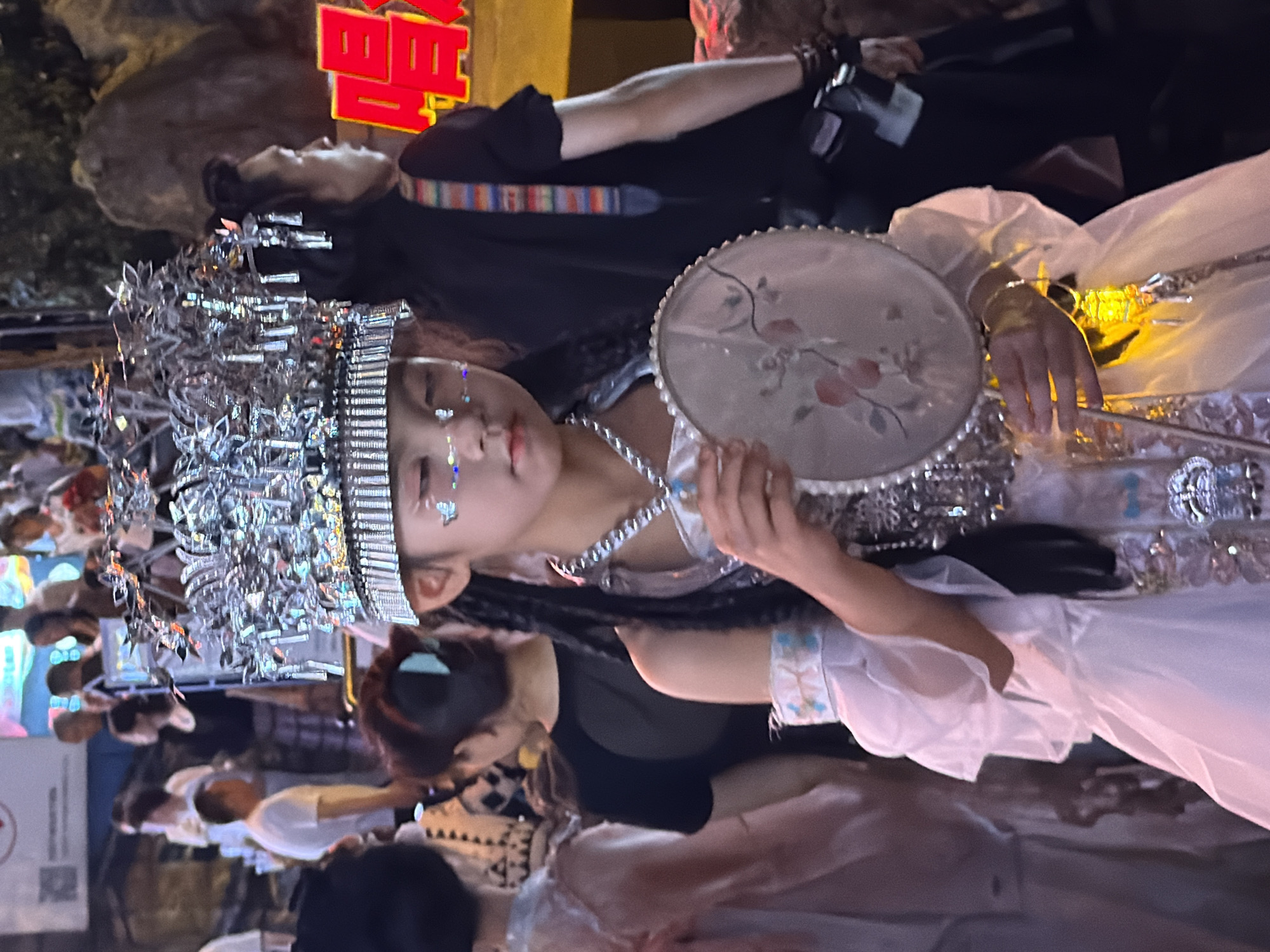A young girl adorned in a white, ornate costume and a metallic headdress stands in the foreground, holding a circular fan. She has glitter on her face and her hair is in braids. Behind her, several adults are visible, some blurred, in what appears to be an outdoor setting at night, possibly a street or market in Yuzhong District, China. In the background, there are illuminated signs and structures, suggesting an urban environment.