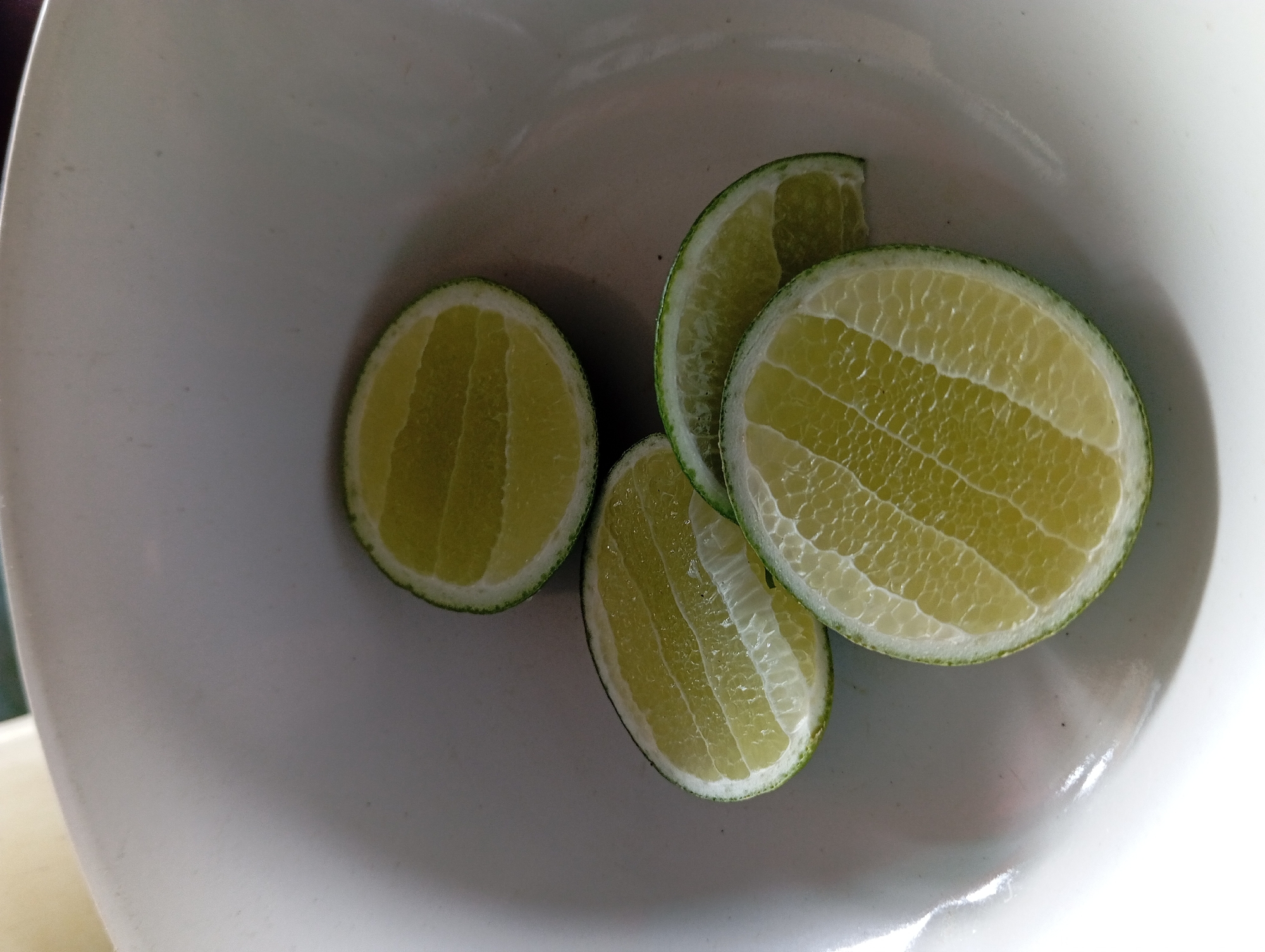 Four slices of lime are arranged in a white bowl. The limes are cut crosswise, revealing their segmented pulp and white pith. The bowl appears to be ceramic and is set against a blurred background. There is no indication of people, actions, or interactions, and no specific location in Lemahbang, Indonesia is discernible from the image.