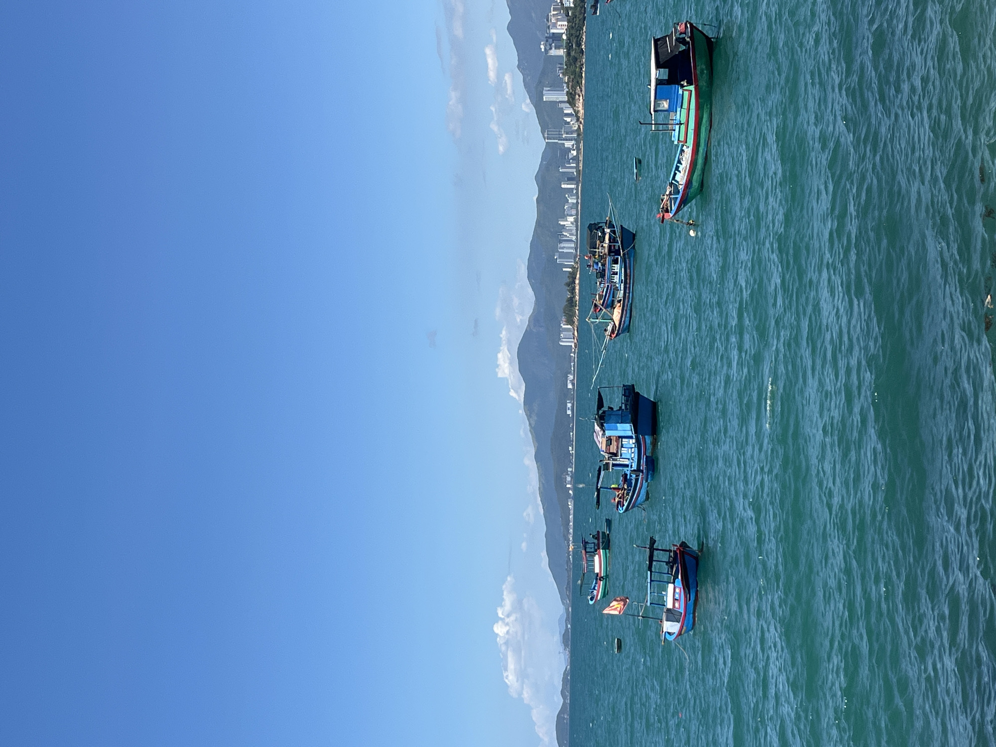 This aerial image shows several boats anchored in the waters off the city of Bac Nha Trang Ward, Vietnam. The scene is dominated by the turquoise-colored ocean and a clear blue sky with a few clouds near the coastline. The coastline itself is visible, featuring a mix of urban development with tall buildings and a mountainous backdrop. Several fishing boats of various sizes and colors are scattered across the water.
