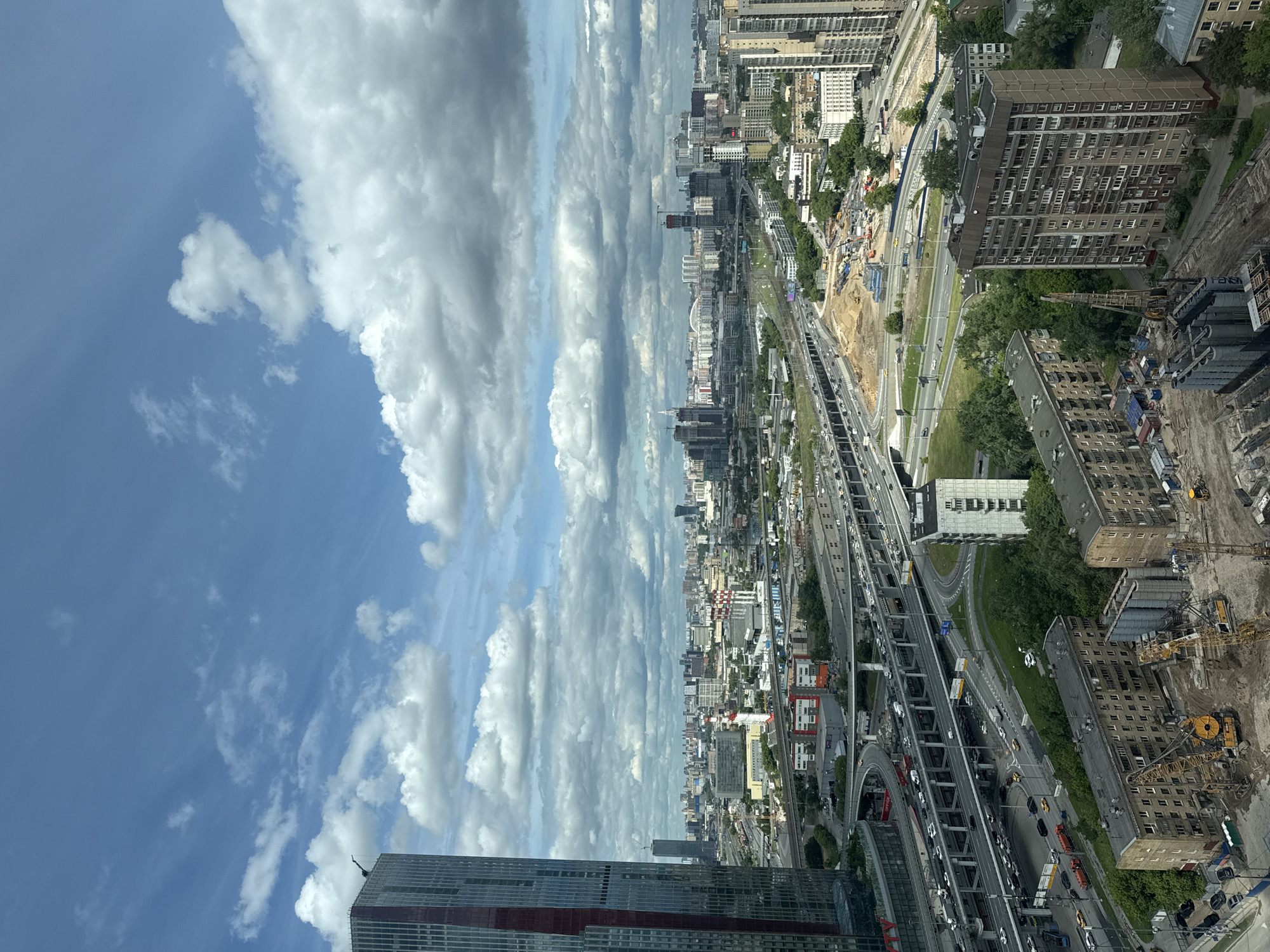 This image shows an aerial view of a city with a prominent highway system cutting through the urban landscape. High-rise buildings are clustered in the distance, while smaller buildings and residential structures dot the area closer to the highway. A significant amount of construction is visible, with cranes and construction vehicles working on what appears to be a new building or infrastructure project. The sky is filled with cumulus clouds and patches of blue sky.
Based on the architectural style, the presence of a large highway, and the urban density, this scene depicts a city in Eastern Europe, potentially in Russia. Given the information provided the location is Khimki, Russia.