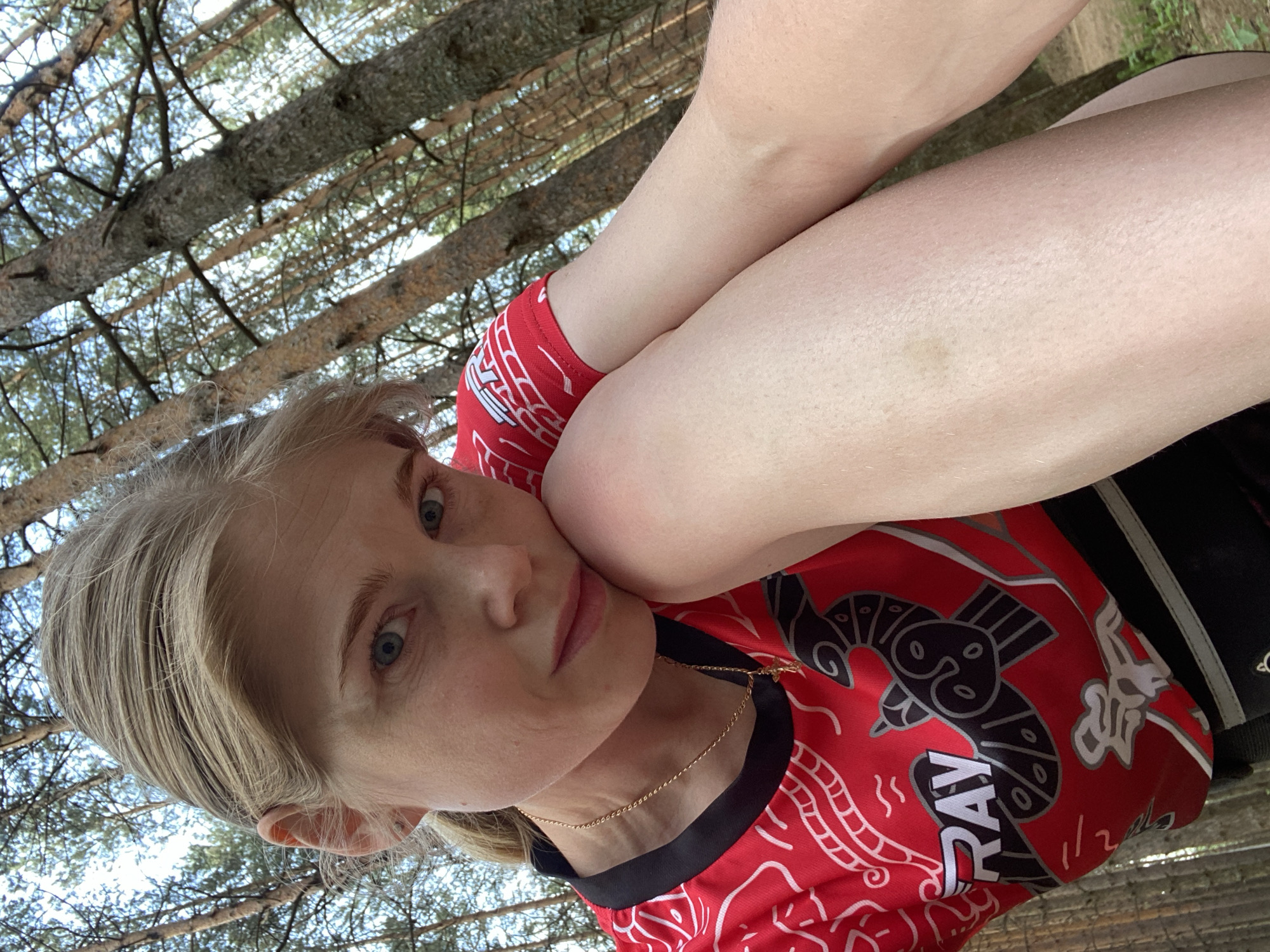 A woman with blonde hair and blue eyes is the main subject of the image. She is wearing a red shirt with black accents and a gold necklace. Her arms are crossed, with one arm resting against her cheek. The background shows a forest with tall trees and glimpses of the sky. The image appears to be taken outdoors, possibly in a park or wooded area. Given the mention of Chaikovsky, Russia, it is reasonable to assume the location is within or near this city. The photograph has an upward angle, suggesting the camera was positioned below the subject.