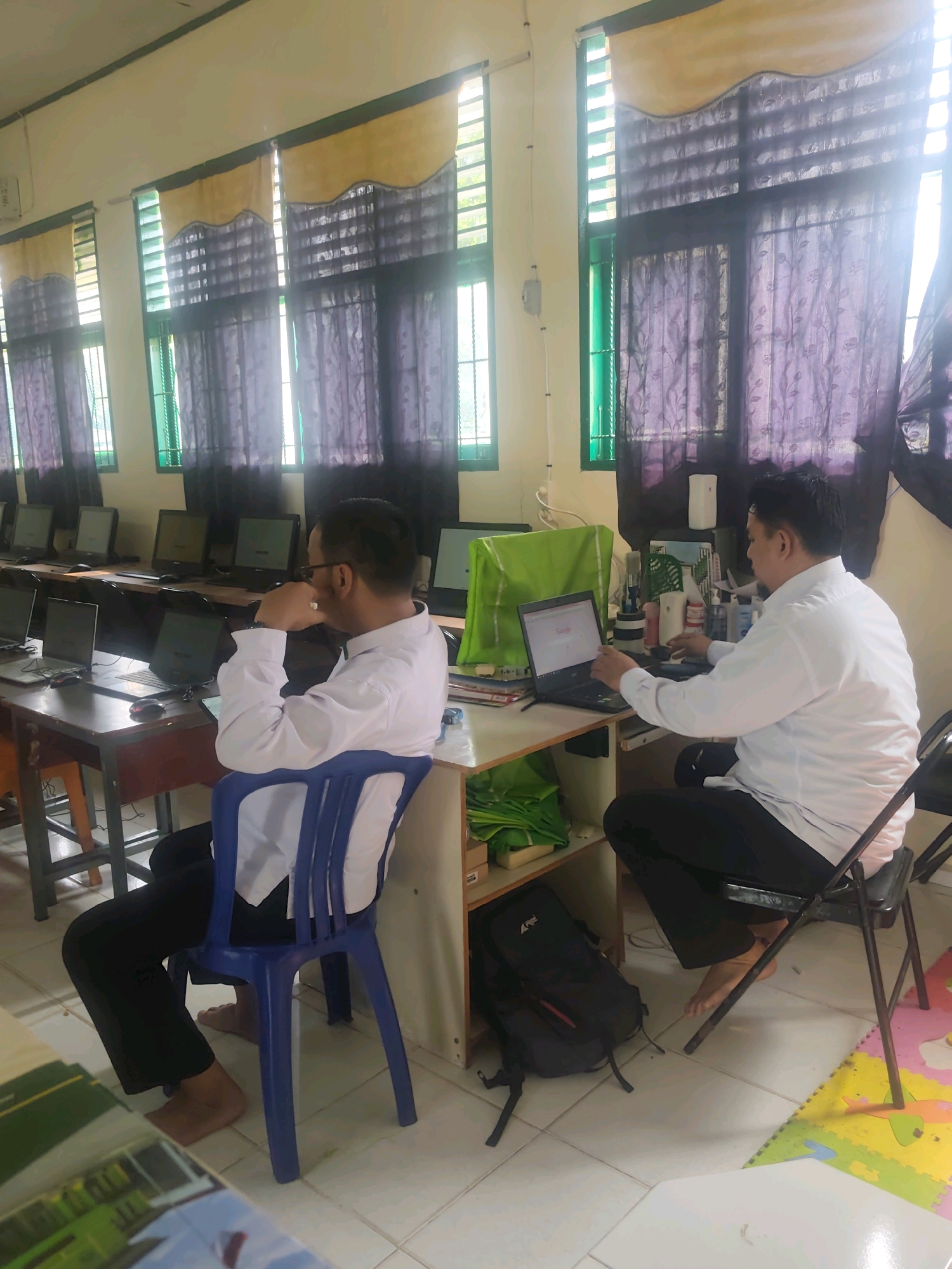 In a classroom in Pematang Reba, Indonesia, two men are seated at desks in front of computers. The room has a tile floor and large windows covered by patterned curtains. The man on the left is wearing a white shirt and dark pants and is seated on a blue plastic chair. He is looking at a laptop on his desk, which is also cluttered with various items. The man on the right is also wearing a white shirt and dark pants and is seated on a black folding chair. He is also looking at a laptop, and appears to be working or studying. In the background, there are several other computer stations with monitors. There are also backpacks and other personal items on the floor and on desks.