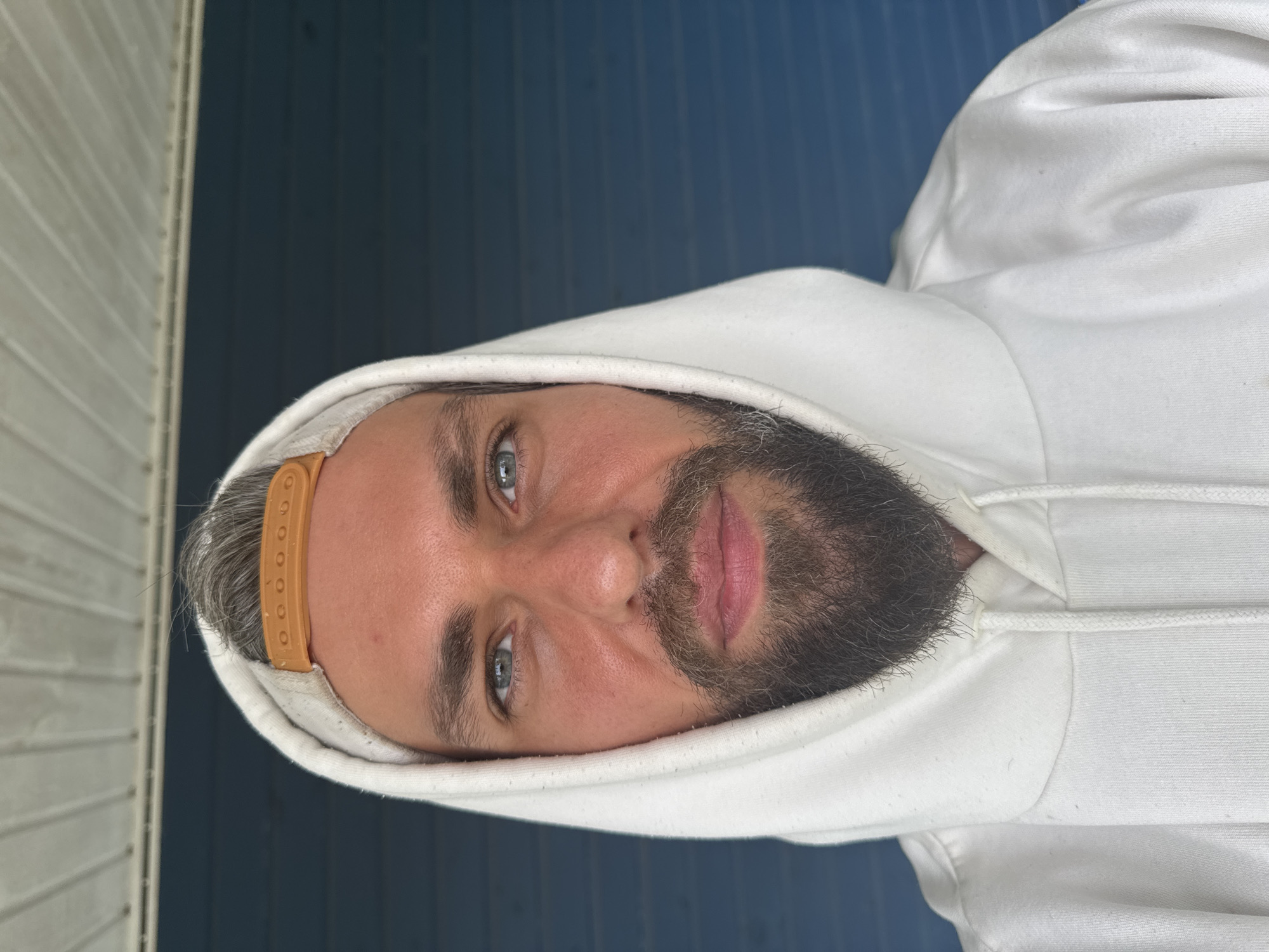 A close-up, head-and-shoulders shot shows a Caucasian man with fair skin, blue eyes, and a beard, wearing a white hooded sweatshirt and a tan baseball cap. The man is looking directly at the camera. The background consists of a dark blue, horizontally paneled surface, and a white, slatted ceiling. The lighting is bright, suggesting an outdoor setting during the day. No specific location in Kuti, Russia, can be identified from the image.