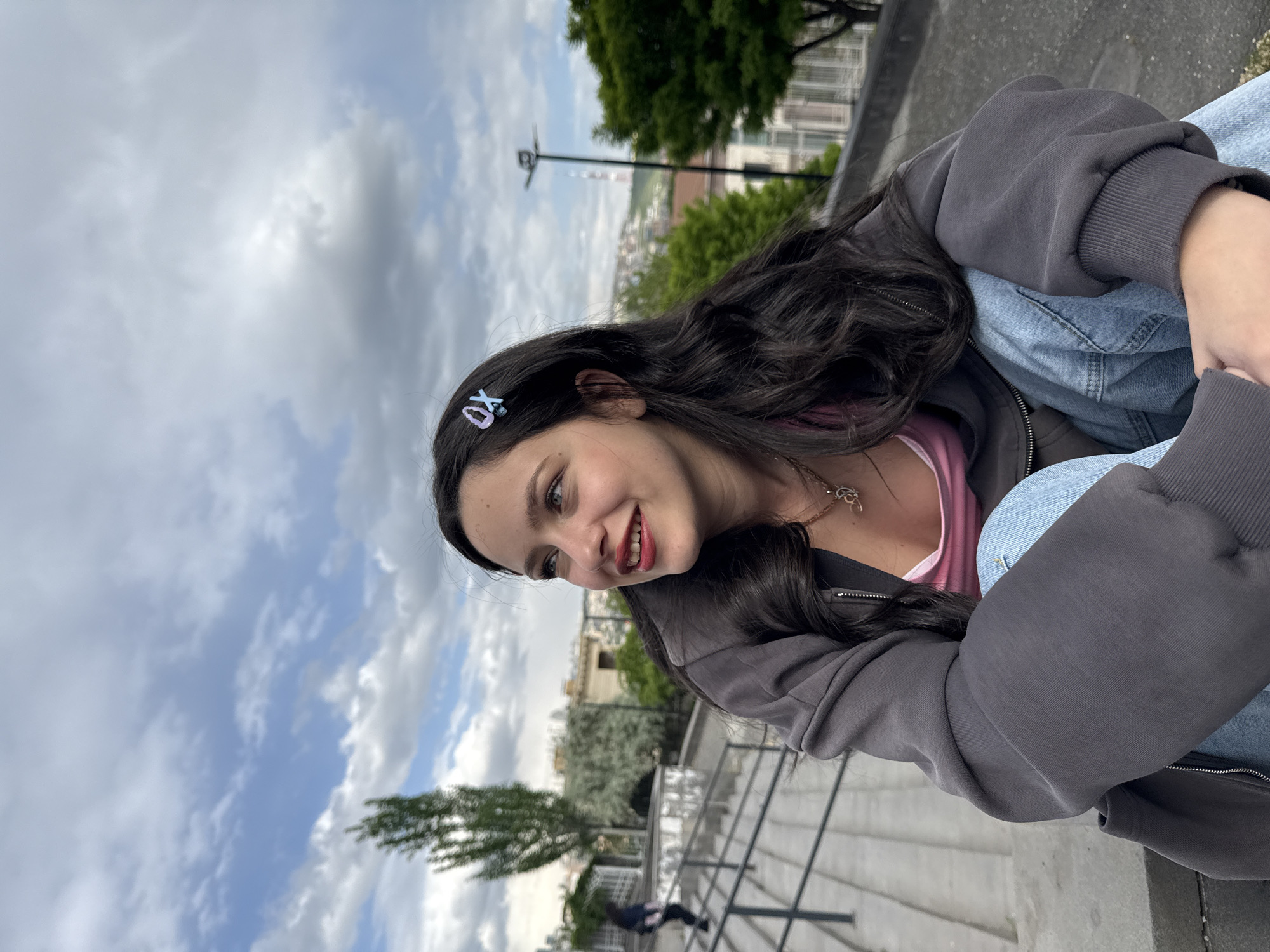 A young woman sits with her legs crossed, arms hugging her knees, on a light gray concrete structure in Yerevan, Armenia. She wears a gray zip-up hoodie, a pink top, and blue jeans, and a blue "XO" hair clip. Her hair is dark and wavy. She is looking to her left with a slight smile. The background includes trees, buildings, and a cloudy sky. A person walks on stairs in the background.