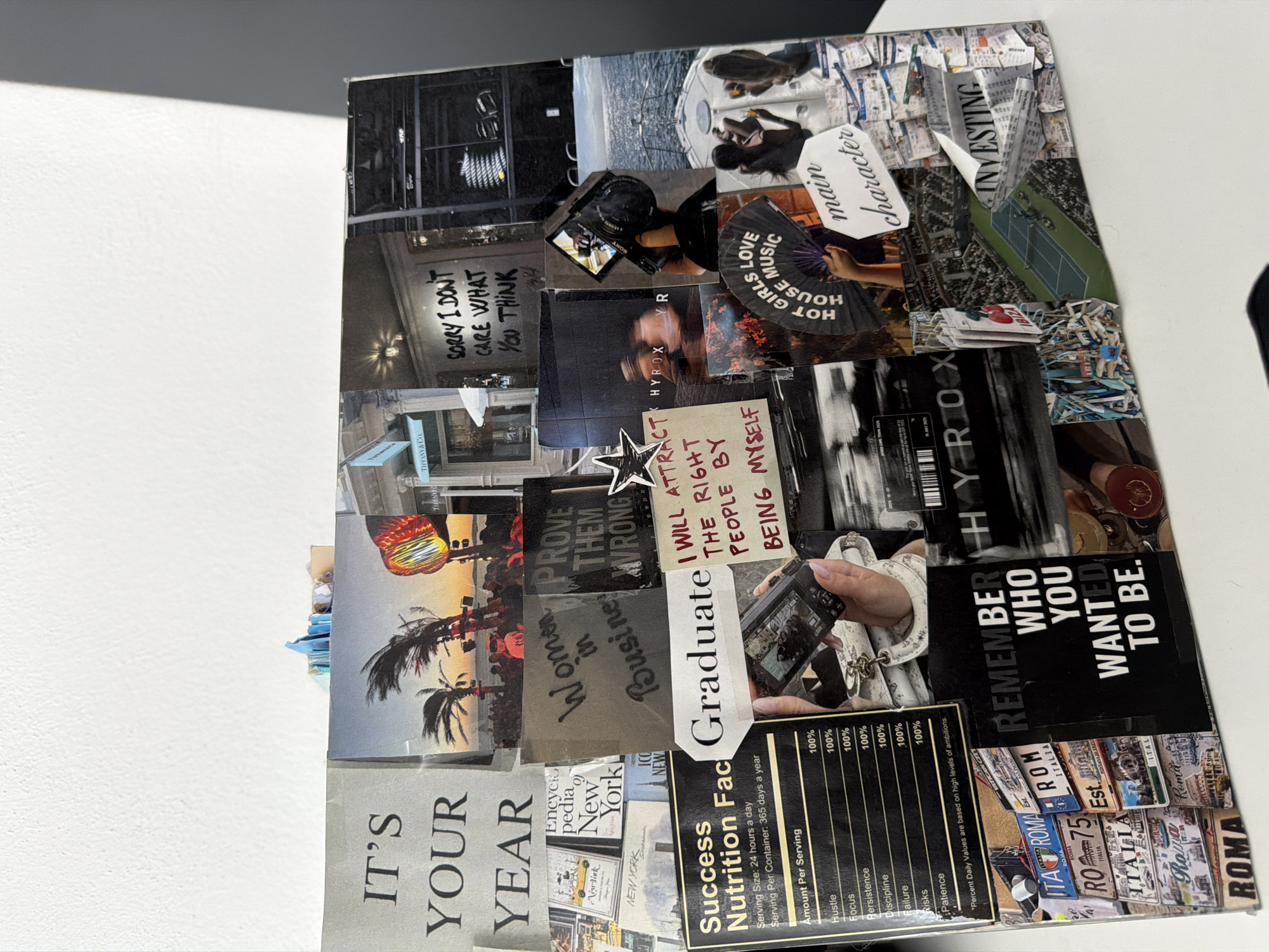 The image shows a collage on a flat surface, possibly a table. The collage consists of various cut-out images and text fragments arranged to form a vision board. Text elements include phrases such as "IT'S YOUR YEAR," "Women in Business," "I WILL ATTRACT THE RIGHT PEOPLE BY BEING MYSELF," "REMEMBER WHO YOU WANTED TO BE," "main character," and "INVESTING." Images feature scenes of urban life, travel destinations (including Rome), and motivational themes. A "Nutrition Facts" label focusing on personal success metrics like "Hustle," "Focus," and "Patience" is prominently displayed. There is no indication of the location being in the city of Puebla, Mexico.