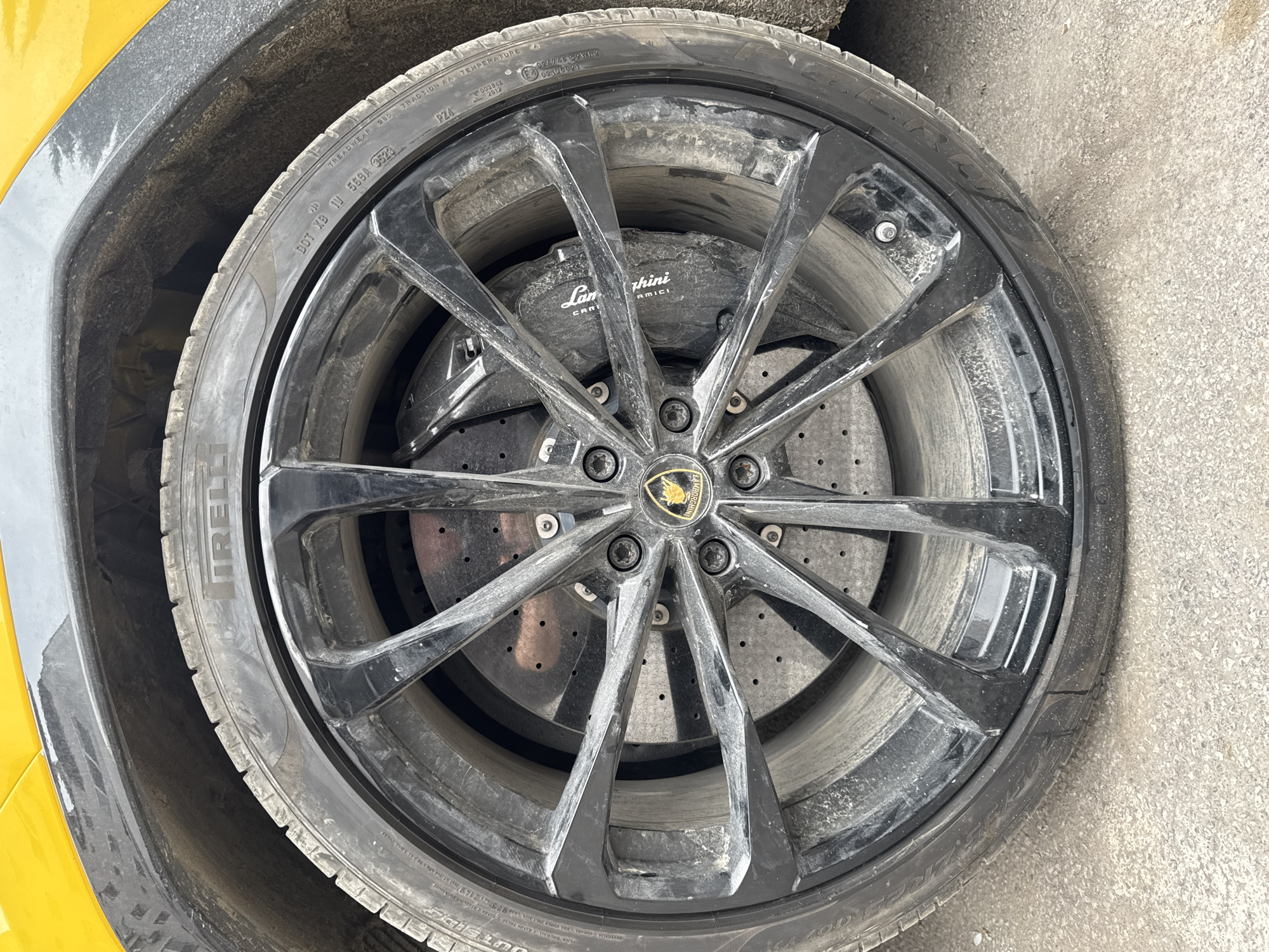 A close-up, eye-level shot captures a dirty black multi-spoke wheel and tire belonging to a yellow Lamborghini, set against a grey, textured ground in Plovdiv, Bulgaria. The wheel is covered in a significant layer of dust and grime, particularly on the glossy black spokes and the carbon-ceramic brake disc, which is visible through the spokes.
The tire is a Pirelli P Zero (PZ4), with "IRELLI" and "PZ4" clearly visible on the sidewall. Other tire markings include "TREADWEAR 260 TRACTION AA TEMPERATURE A" and the DOT code "XB TV 569A 3523", indicating manufacture in the 35th week of 2023. Behind the spokes, a large black brake caliper is prominent, featuring the "Lamborghini" logo and the word "CERAMICI," signifying carbon-ceramic brakes. The brake disc itself is perforated. The center of the wheel proudly displays the iconic Lamborghini bull logo on its cap.
A portion of the car's bright yellow fender is visible on the left side of the frame, contrasting with a grey plastic wheel arch liner. The ground beneath the tire is a rough, grey surface, likely concrete or asphalt, also bearing some dirt and debris. The lighting suggests a bright, overcast day, as there are no harsh shadows. The overall impression is of a high-performance vehicle that has been driven and is currently somewhat soiled, rather than pristine.