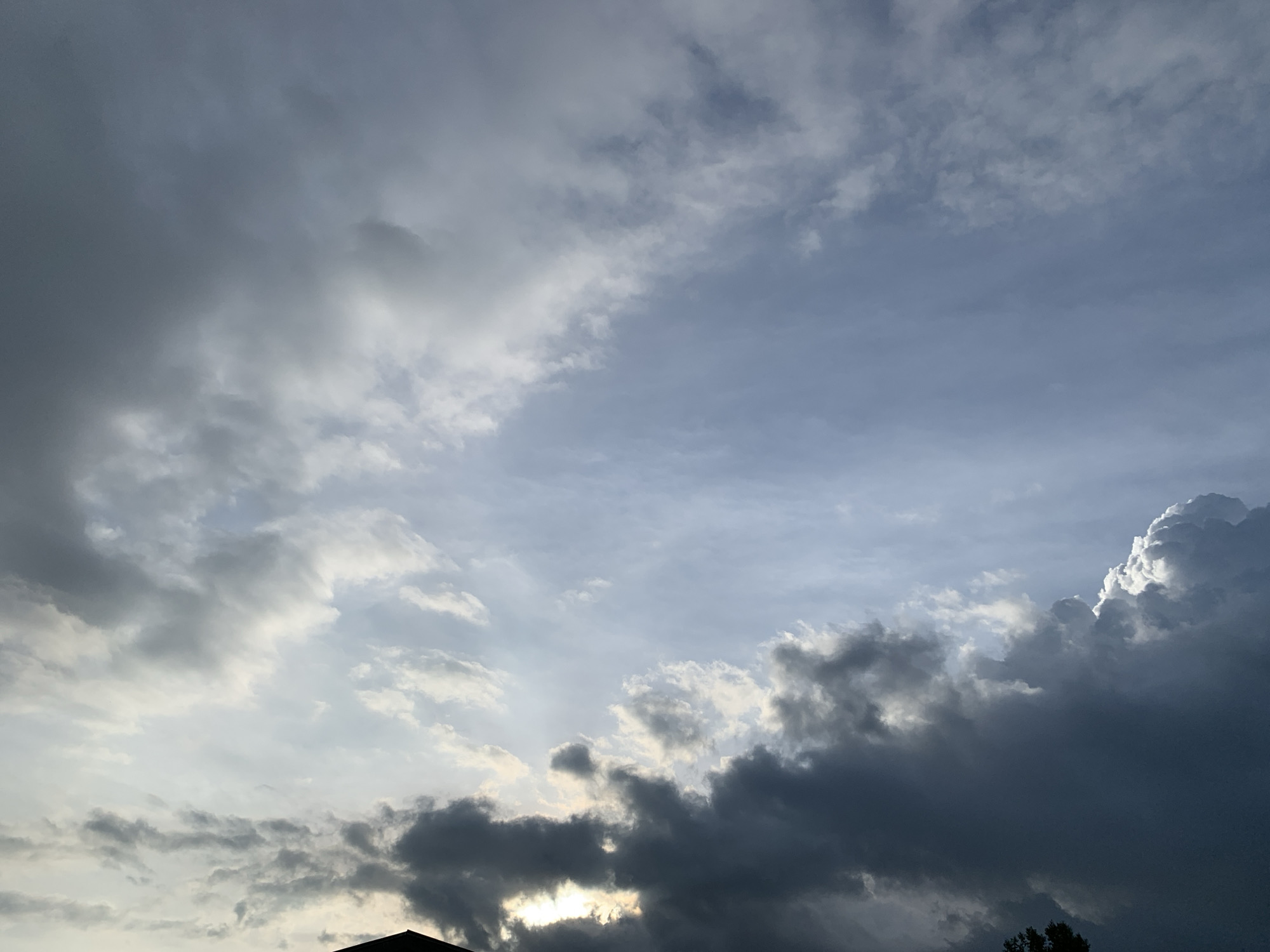 The image depicts a dramatic sky filled with a variety of clouds, suggesting an impending storm or a recent passing of one. The clouds range in color from dark, heavy grays to lighter, wispy whites, with pockets of bright sunlight breaking through. This interplay of light and shadow creates a sense of depth and movement across the sky. The scene appears to be a wide, open sky, possibly an aerial view or taken from a high vantage point, as there are faint silhouettes of rooftops and treetops at the very bottom edge of the frame. The presence of these earthly elements grounds the otherwise ethereal cloudscape. The atmosphere is charged, hinting at meteorological activity, but there are no visible people or specific actions taking place. The location within Kuru Karama, Nigeria, adds a geographical context to the natural phenomenon.