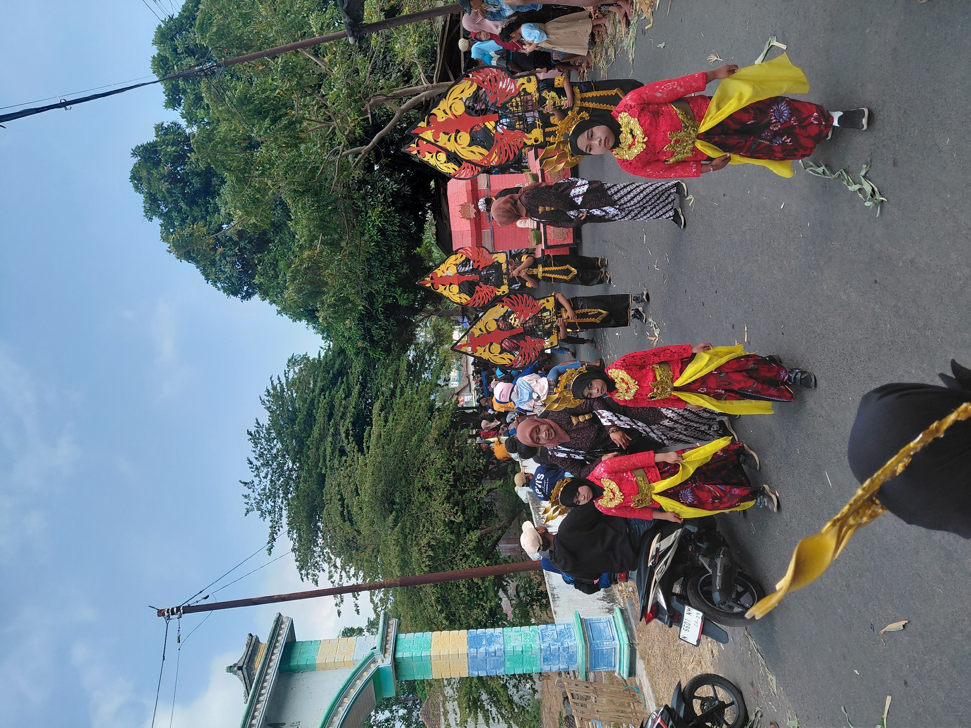 This is a photograph taken from ground level, looking slightly upwards, on a paved street in Bimo, Indonesia. The sky is a pale blue with some wispy clouds. In the foreground, a person wearing a black hijab and a gold, decorative item is partially visible, holding it up.
To the right of the frame, four young girls are walking, dressed in traditional Indonesian attire. They wear bright red tops with intricate gold embroidery and embellishments around the neckline, and matching red skirts with a floral pattern, adorned with yellow sashes. Their hair is covered by dark hijabs, and one girl wears a small gold crown.
Behind the girls, a procession is taking place. Several individuals are holding large, colorful puppets, which appear to be traditional Wayang Kulit figures, with red, yellow, and black designs. Some adults in the procession are also wearing traditional clothing, including batik patterns.
On the left side of the image, a decorated archway with blue, yellow, and white painted sections stands prominently. A motorcycle is parked to the left of the archway, and a few people are gathered around it. In the background, lush green trees line the street, and the silhouette of buildings and more people can be seen. The overall scene depicts a cultural event or parade.