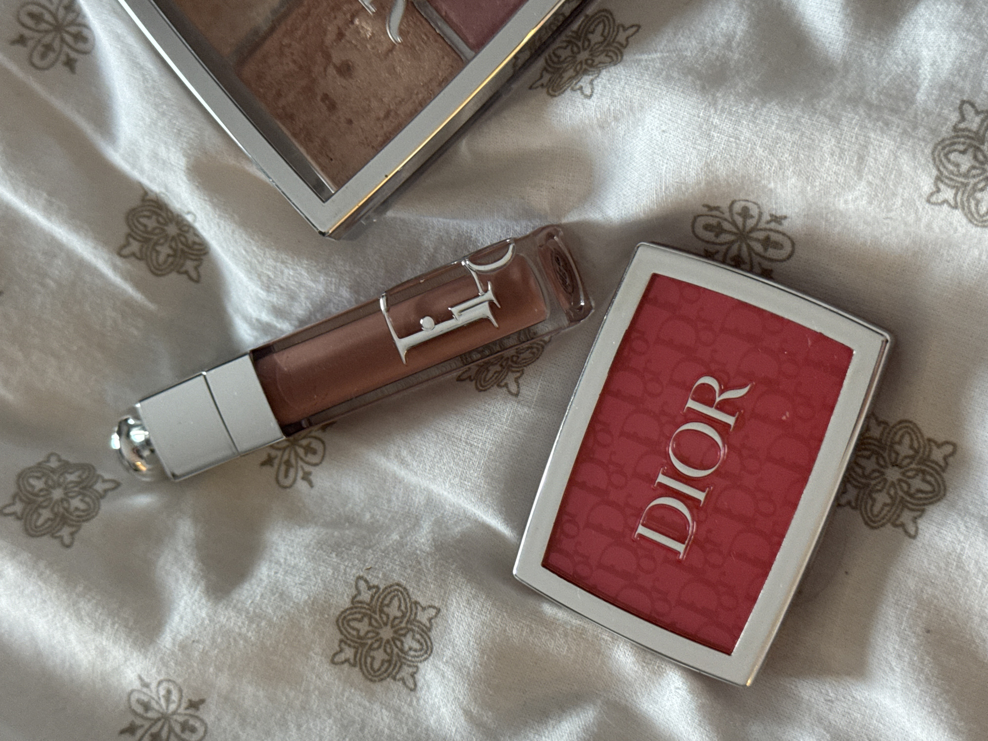 The image shows an overhead shot of three cosmetic items on a white fabric with a repeating floral pattern. The objects are positioned relatively close together, suggesting they are arranged in a casual manner.
The top left item is a makeup palette, possibly containing blush or bronzer shades, encased in a silver rectangular container. The second item is a clear tube of lip gloss or lipstick with a silver cap. The Dior logo is clearly visible on the tube. The third item, located on the right, is a red blush compact also in a silver container with a repeating Dior logo pattern and the brand name DIOR embossed in white.
The backdrop fabric has light variations and shadowing, providing depth to the image.
The location and city where the image was taken are not specified.