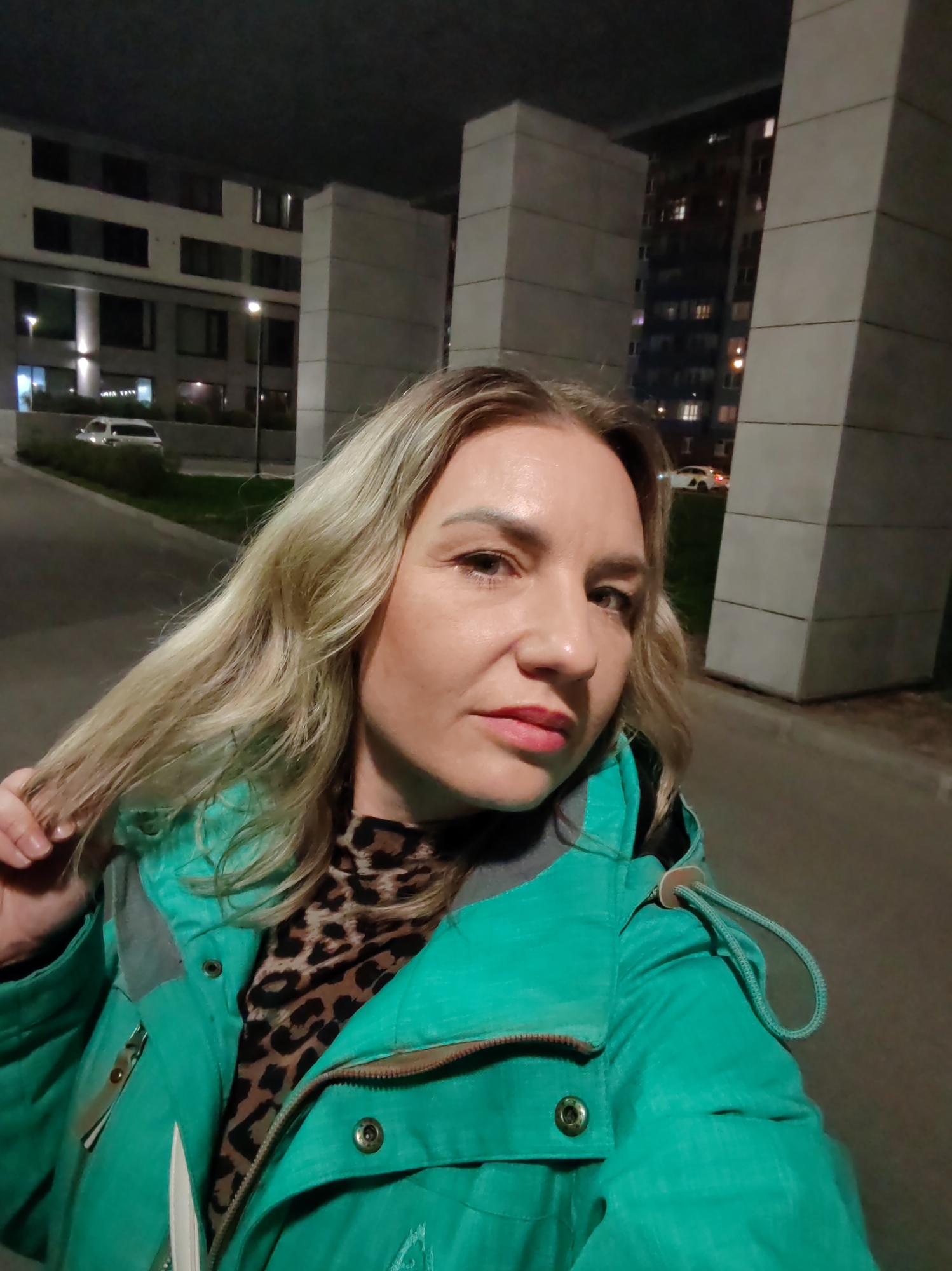 A woman with light blonde hair is holding a strand of her hair with her left hand and looking toward the camera. She is wearing a bright green winter jacket with a leopard print top underneath. The background shows the urban environment in Saint Petersburg, Russia at night with several buildings, lights, a road and greenery. Several vehicles are parked on the side of the road.