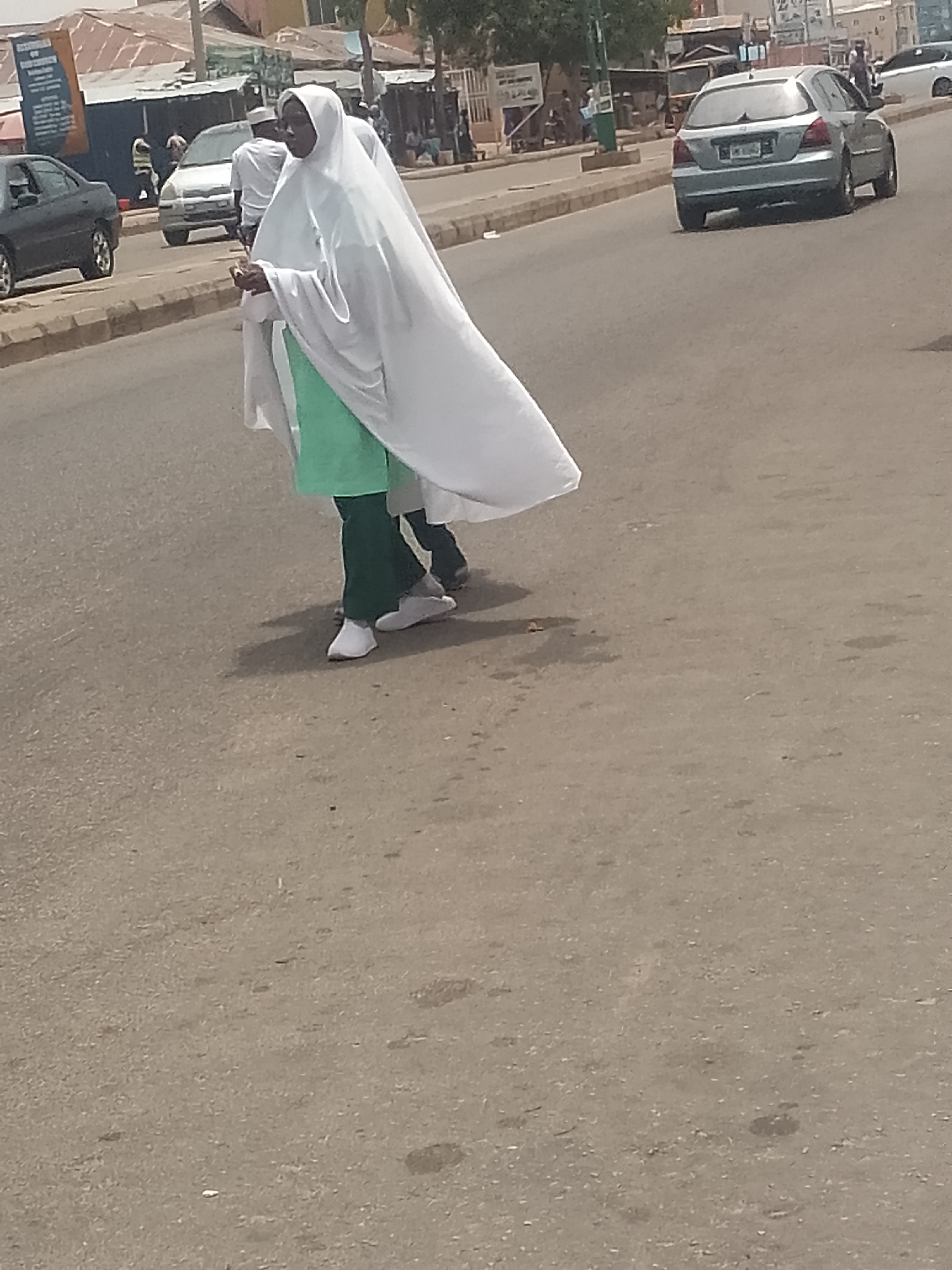 Two women in white hijab and green uniforms walk across a paved road. A silver car drives on the road behind them, heading away from the camera. Other cars are visible further away. Buildings and shops line the street. The image appears to be taken during the day in Pantami, Nigeria.