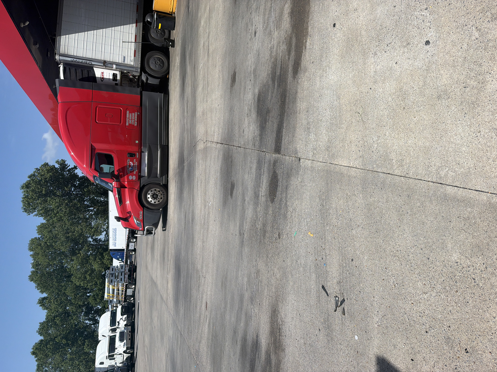 The image captures an outdoor scene in Hayti, United States of America, likely a truck stop or industrial yard, on a sunny day under a clear blue sky. In the foreground, a concrete surface shows signs of wear and tear with oil stains and scattered debris, including what appears to be nail clippers, a bottle cap, and small pieces of colored plastic.
Dominating the mid-ground is a large, red semi-truck parked next to a red overhang structure, possibly a canopy at a fueling station. The truck has the number "2649" visible on its side, and branding that includes "HOMELAND" and "TRAILER". A white refrigerated trailer is attached to the truck, displaying the number "52540" and "CALVIL". Two dark-colored trash receptacles, one black and one yellow, are situated near the rear of the truck.
In the background, several other white semi-trucks are parked, some with trailers. One trailer displays the text "J.R. SCHUGEL". A line of mature, leafy green trees forms a backdrop behind the trucks. The overall impression is one of a functional service area for commercial vehicles.