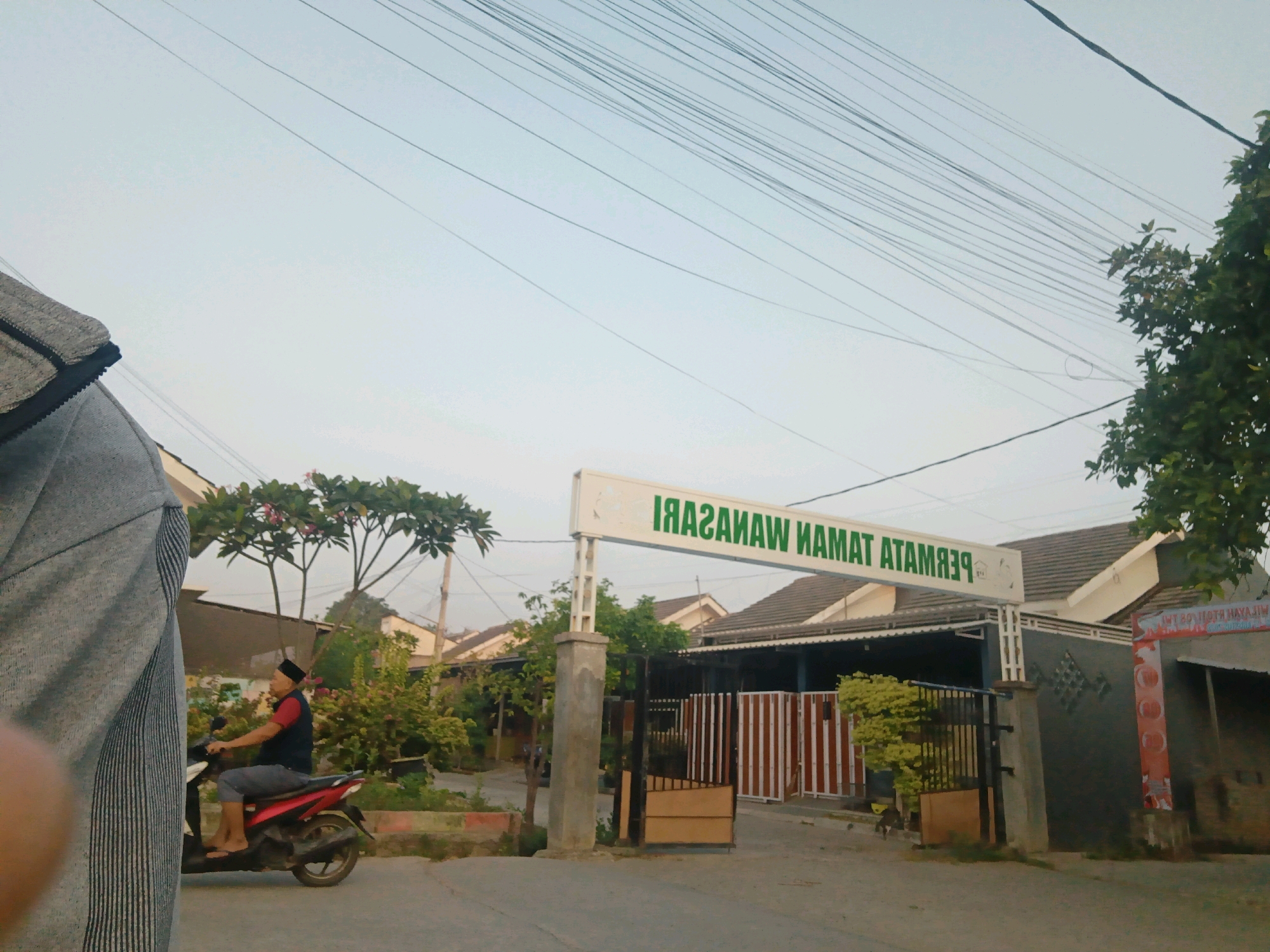 In the city of Wanasari, Indonesia, a street scene unfolds with a low-angle shot. The sky is a pale blue, crisscrossed with numerous power lines. In the foreground, a portion of a person's torso and shoulder, wearing a grey and black patterned garment, is visible on the left side of the frame. To the right, a man wearing a black cap, a red shirt, and a dark vest, is riding a red motorcycle. He is facing left, with his feet resting on the footpegs and his left hand on the handlebars. The motorcycle is parked on a paved road. Behind the motorcycle, a residential area is apparent, with houses featuring tiled roofs and various foliage. A prominent sign with green lettering on a white background reads "TAMAN MAMA WANASARI ATAMBA," indicating a location, likely a housing complex or park. The sign is supported by concrete pillars. A black metal gate with vertical bars and a wooden section is visible in front of the entrance to the residential area. Further to the right, a vertical red and white banner is attached to a building. The overall scene suggests a typical residential street in Indonesia.