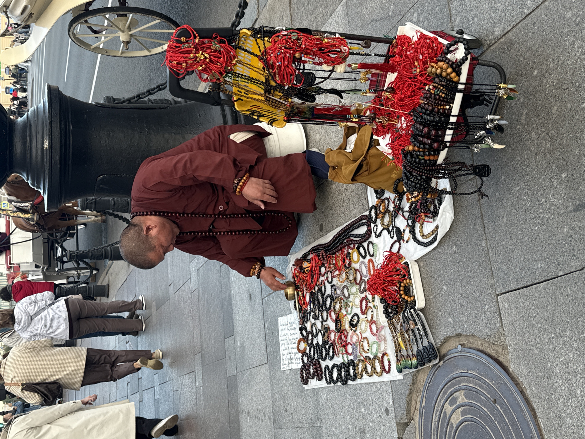 The image captures a street scene in Saint Petersburg, Russia, specifically likely on Nevsky Prospekt, a prominent thoroughfare. In the foreground, a man wearing a maroon robe and dark prayer beads is crouched down, tending to a display of bracelets and necklaces laid out on a white cloth. The items for sale are primarily made of wood and stone beads, with some featuring red string and small charms. Beside him, a small cart is laden with more jewelry, predominantly red strings and dark beads. The man is interacting with a small brass bowl placed among the merchandise. In the background, several people are walking by, and a horse-drawn carriage, a common sight for tourists in the city, is visible. The ground is paved with granite cobblestones. The overall ambiance suggests a busy urban environment where street vendors offer their wares to passersby.