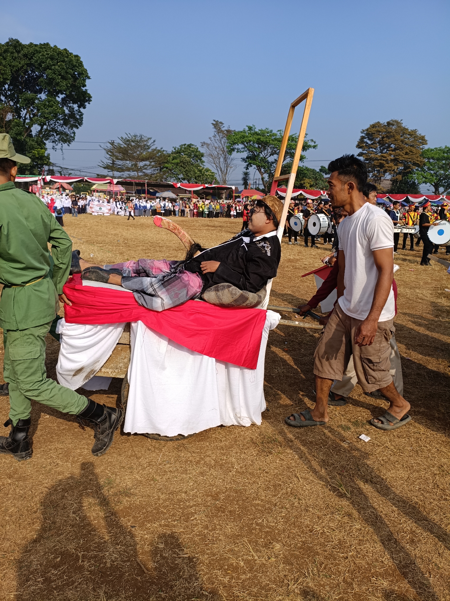 A group of men participate in a parade in Cibodas, Indonesia. In the foreground, a man in a green uniform walks next to a wooden structure on wheels, draped in red and white fabric. A second man, wearing traditional Indonesian attire, lies on the structure, his head tilted back. To the right, another man in a white t-shirt and khaki shorts walks beside the structure. In the background, a marching band with drums can be seen, as well as a crowd of spectators gathered behind tents and banners. The scene unfolds outdoors on a dry, grassy field under a clear blue sky.