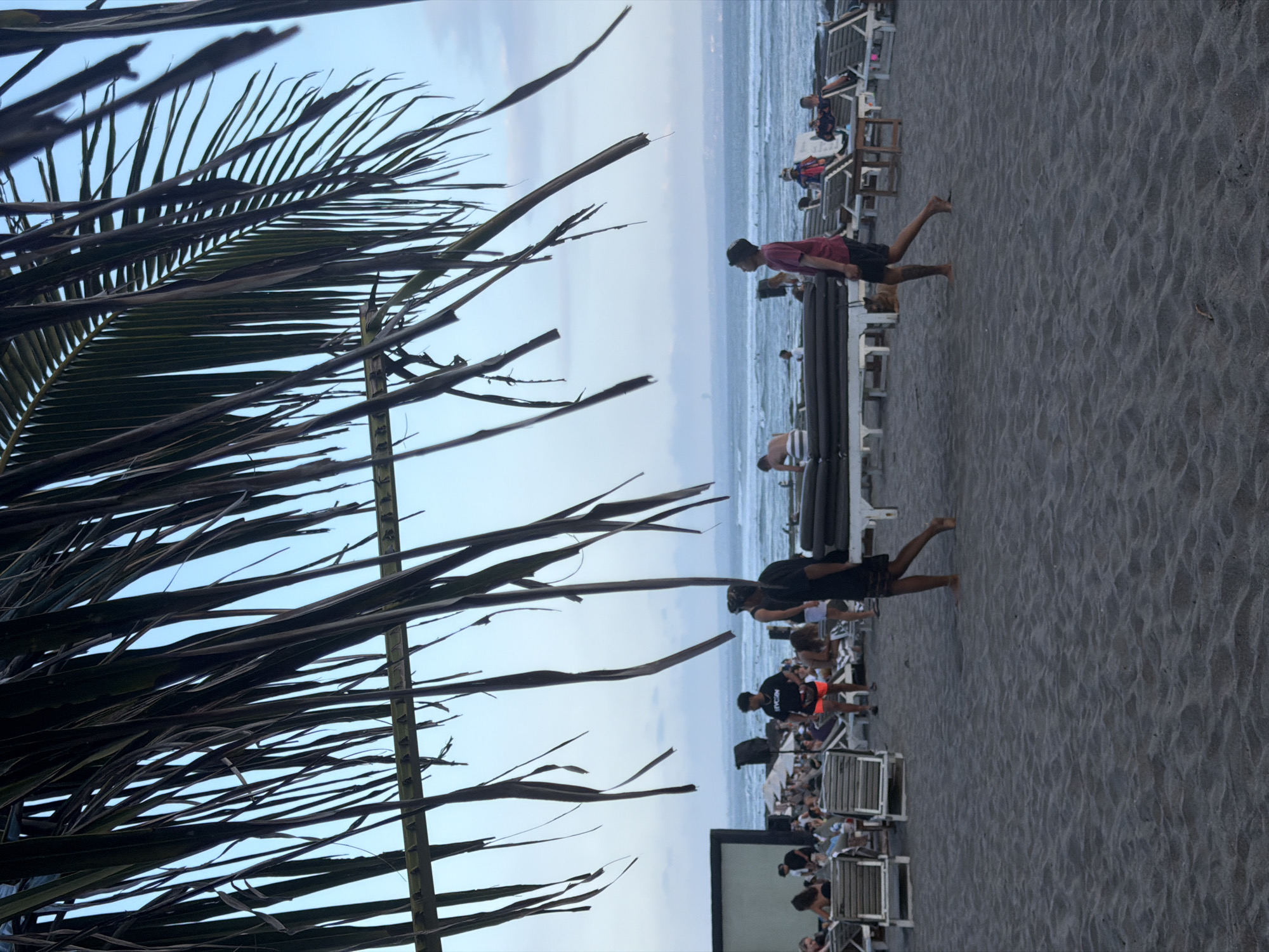 The image depicts a beach scene in North Kuta, Indonesia. In the foreground, dark sand covers the ground, with the impression of waves or ripples visible. Overhanging palm fronds partially obscure the view of the sky.
In the middle ground, two young men are walking on the sand. The man on the left is wearing a black t-shirt and orange shorts. The man on the right is wearing a red t-shirt and black shorts. Several rows of lounge chairs, some occupied by people, are set up on the sand behind them, facing the ocean. A low structure with dark cushions, possibly a daybed or seating area, is positioned between the lounge chairs and the people. Further back, the ocean stretches to the horizon, with gentle waves and a few distant figures in the water, suggesting people are swimming or surfing. The sky is a pale blue with a few scattered clouds.
In the background, a building or structure is partially visible on the left, with more lounge chairs and people around it. The overall atmosphere suggests a relaxed, daytime beach setting.
