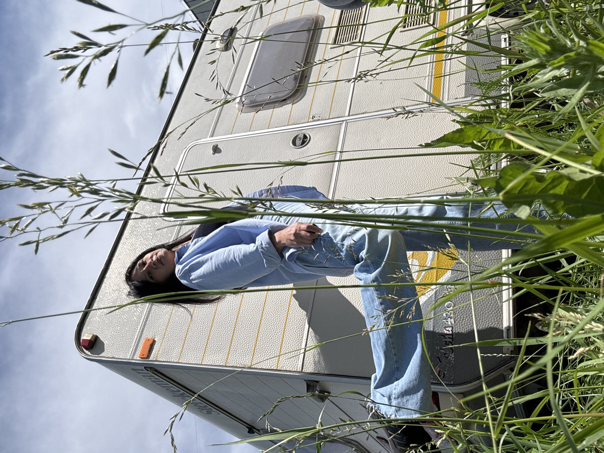 A low-angle shot shows a woman standing in tall grass in front of a white caravan under a partly cloudy sky. She is wearing a light blue collared shirt and light blue jeans, with her left leg bent and extended forward. The caravan has the brand name "Sterckeman" visible on its side and yellow stripes running horizontally. The scene appears to be in an outdoor, possibly rural or semi-rural area. The woman's face is partially obscured by blades of grass, and she is looking upwards. The lighting suggests it is daytime.