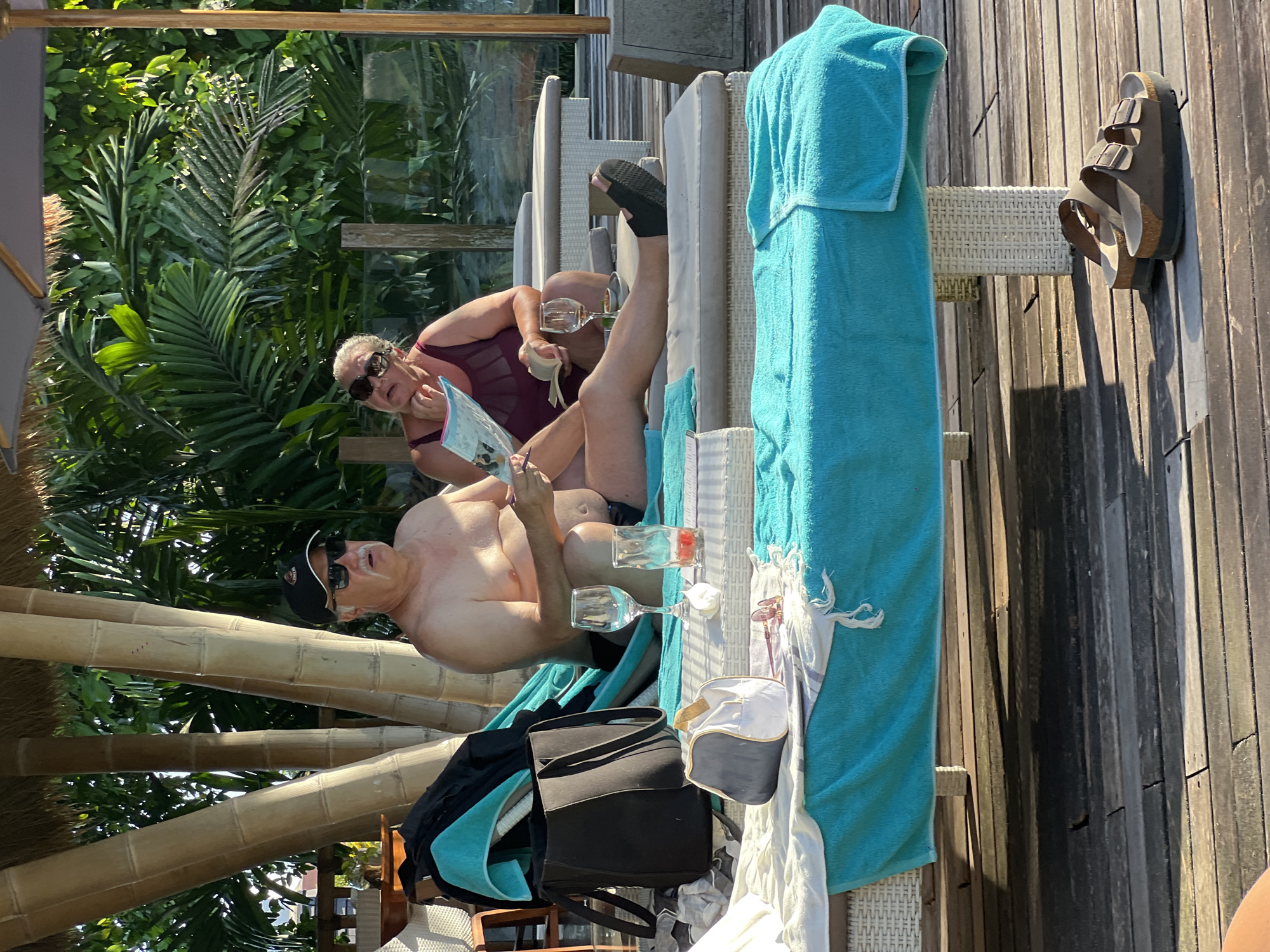 Two people are relaxing on lounge chairs outdoors in North Kuta, Indonesia. One person is wearing a black cap and sunglasses, and they are holding a book. The other person is wearing sunglasses and a maroon swimsuit, and they also appear to be reading. There are wine glasses and a water bottle nearby on the lounge chairs. A blue towel is draped over the chair. A black bag and a white and gray bag are also on the chairs. In the background, there are palm trees and other tropical plants. The lounge chairs are on a wooden deck.