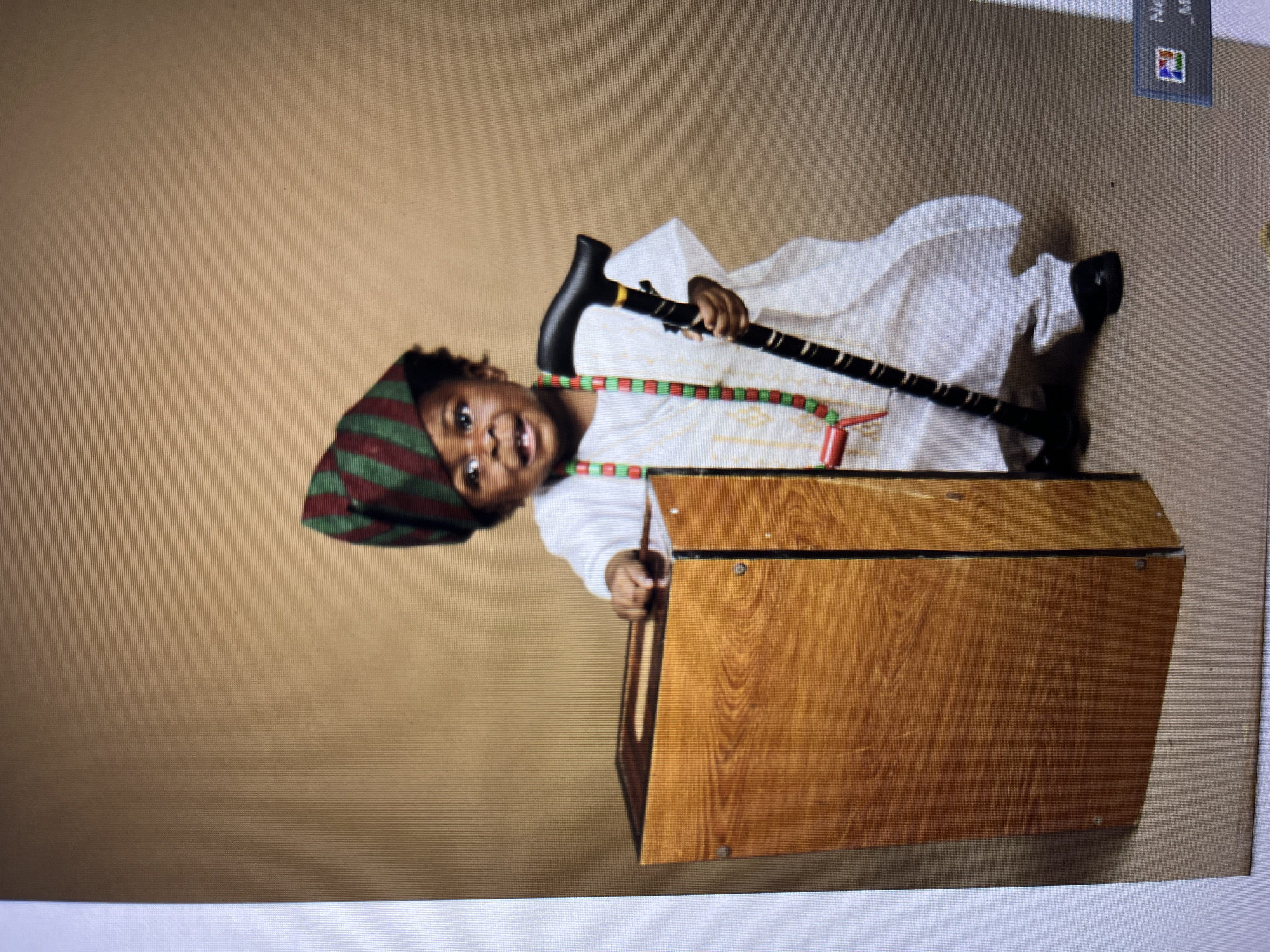 A young child, likely a toddler, is posed for a studio portrait against a plain, tan backdrop. The child is dressed in traditional Nigerian attire, including a white agbada and a striped green and red fila (cap). A beaded necklace with red and green beads is worn around the child's neck. The child is standing and leaning on a wooden box-like object, which appears to be a prop. In their right hand, the child holds a black cane with a curved handle and decorative yellow bands. The child's expression is one of slight surprise or amusement, with an open mouth and visible teeth. The lighting is soft and diffused, characteristic of a studio setting. There is no visible indication of the city of Gura Top, Nigeria, in the image.