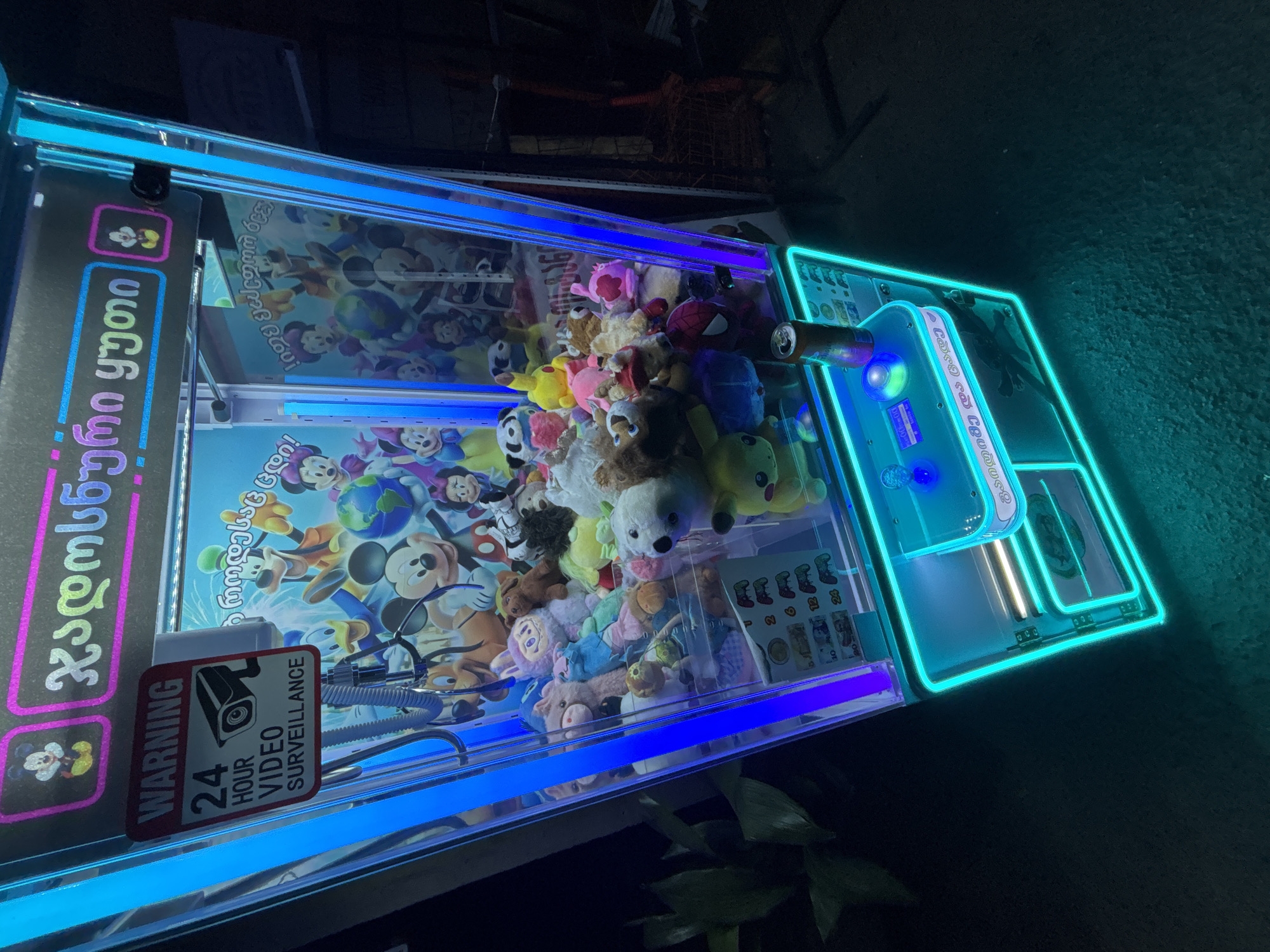 This is a close-up, low-angle outdoor shot of a neon-lit claw machine, likely located in Tbilisi, Georgia, given the Georgian text on its signage. The machine is filled with plush toys, including characters resembling Mickey Mouse and other Disney figures. A prominent "WARNING 24 HOUR VIDEO SURVEILLANCE" sticker is affixed to the left side of the machine. The machine has a coin slot, a joystick, and various buttons for operation. The surrounding environment is dark, with hints of foliage on the left and indistinct structures on the right, suggesting it might be situated in a public space or arcade at night. The overall impression is one of a typical amusement or entertainment machine.