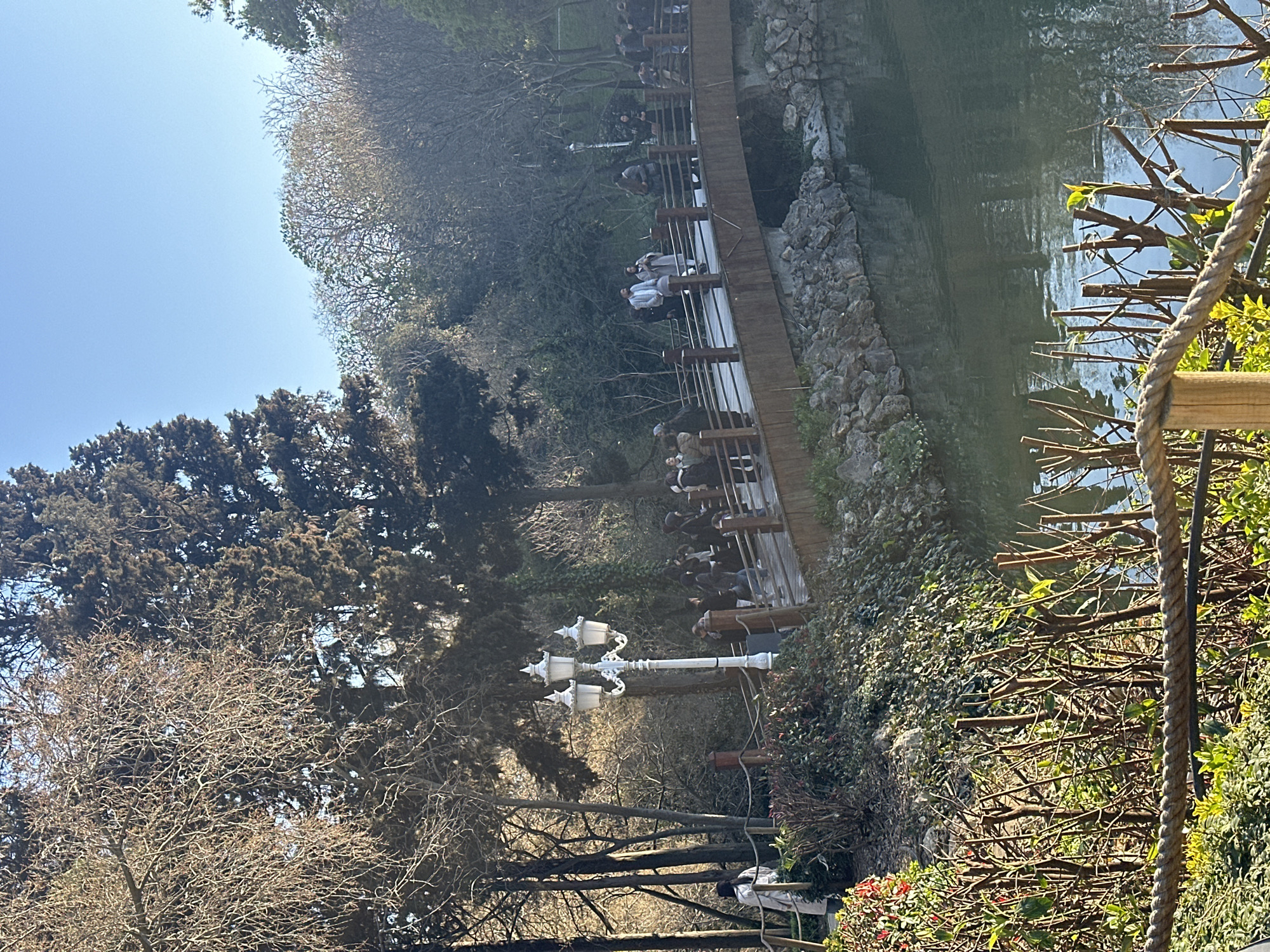 The photograph shows a scene in Beşiktaş, Turkey, likely in a park or garden setting adjacent to water. People are present on a raised wooden walkway, appearing to be walking along or pausing to look at the water. The walkway is lined with wooden posts and a railing on both sides, providing a barrier and likely a viewpoint. The water's edge is reinforced with rough-hewn stones. The foreground exhibits foliage, including branches, plants, and a rope barrier. There is a distinct white streetlamp located in the mid-ground. Several trees with both bare branches and dense foliage populate the scene. The sky is clear and blue.