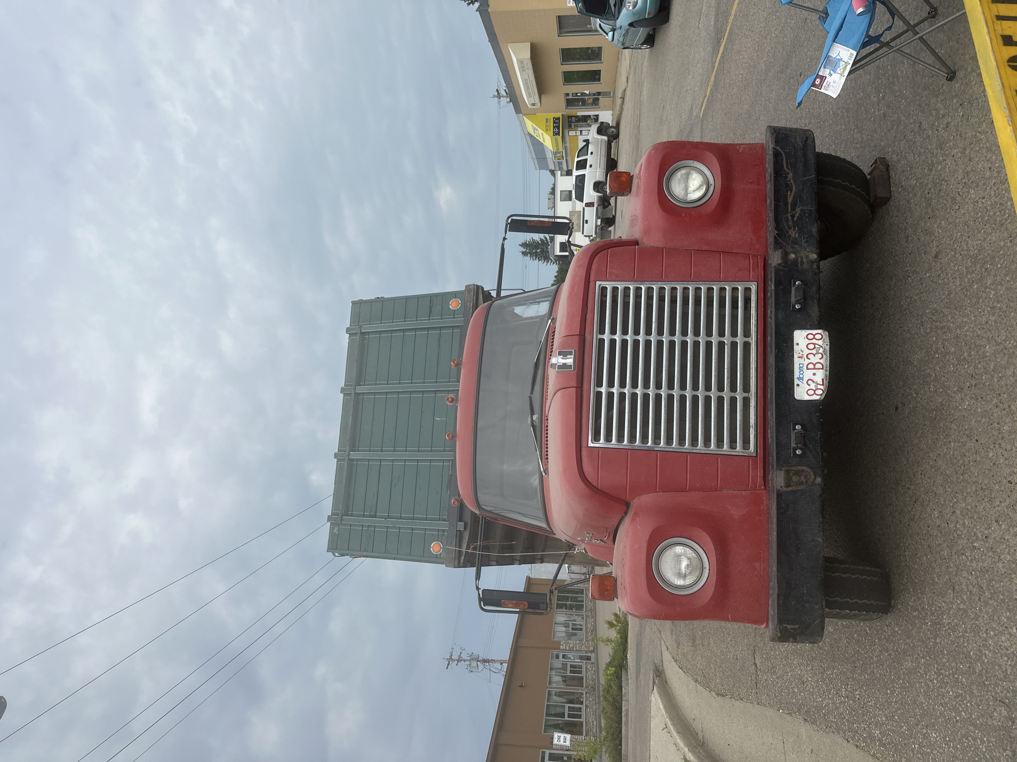 This media file features a close-up, ground-level view of the front of a red truck. The truck has a large, chrome grille and a visible license plate that reads "82 B398 Alberta". The vehicle appears to be parked on an asphalt surface, possibly a street or parking lot, with a curb visible in the foreground. Behind the truck, several buildings are visible, suggesting an urban or commercial setting. One building has a yellow facade with windows and signage, and another is a lighter color with stone accents and windows. The sky is overcast with scattered clouds. There is a folding blue chair with a pink cup on it to the right of the truck. In the background, other vehicles, including a white pickup truck and a light blue car, are parked. The overall scene is daytime. The angle of the shot makes the truck appear dominant in the frame.